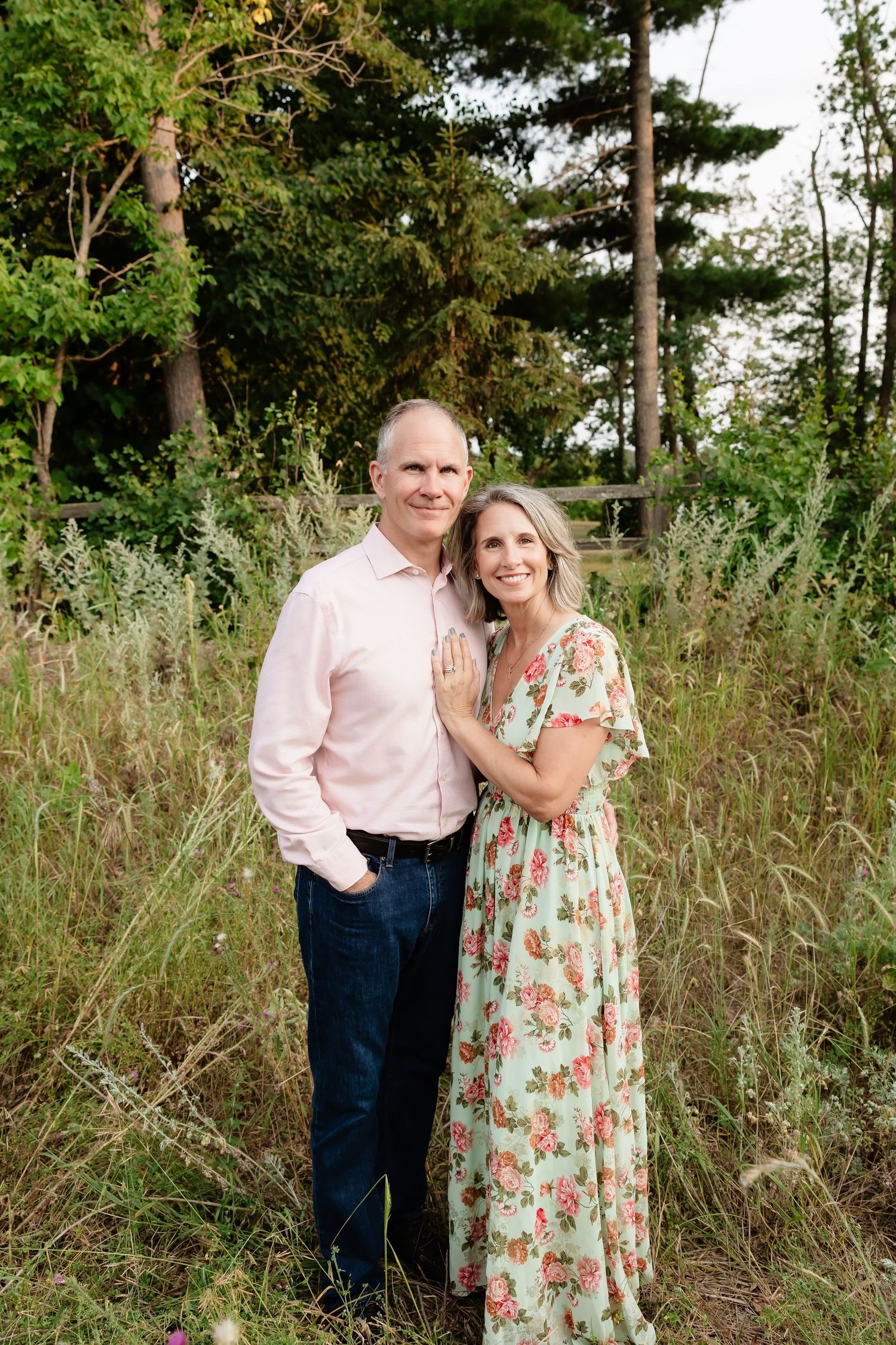 A smiling couple poses outdoors in a lush green field, man in pink shirt and woman in floral maxi dress.