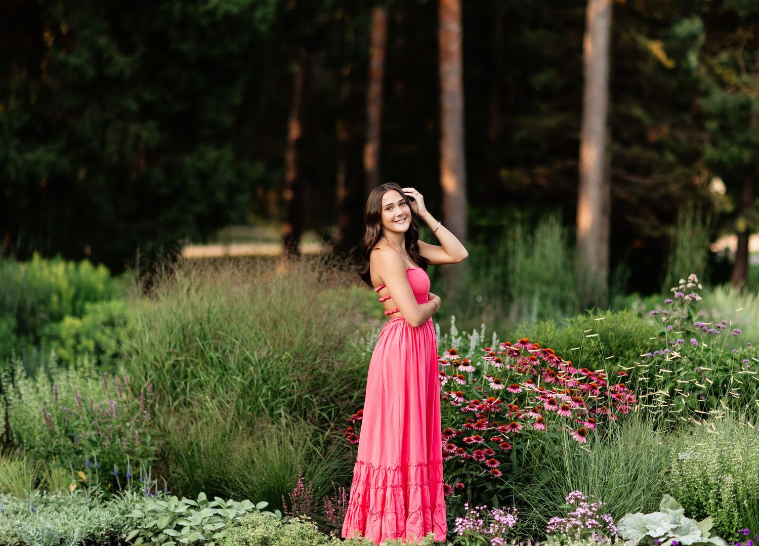 Young woman in pink maxi dress posing in lush garden with colorful flowers and tall trees in background.