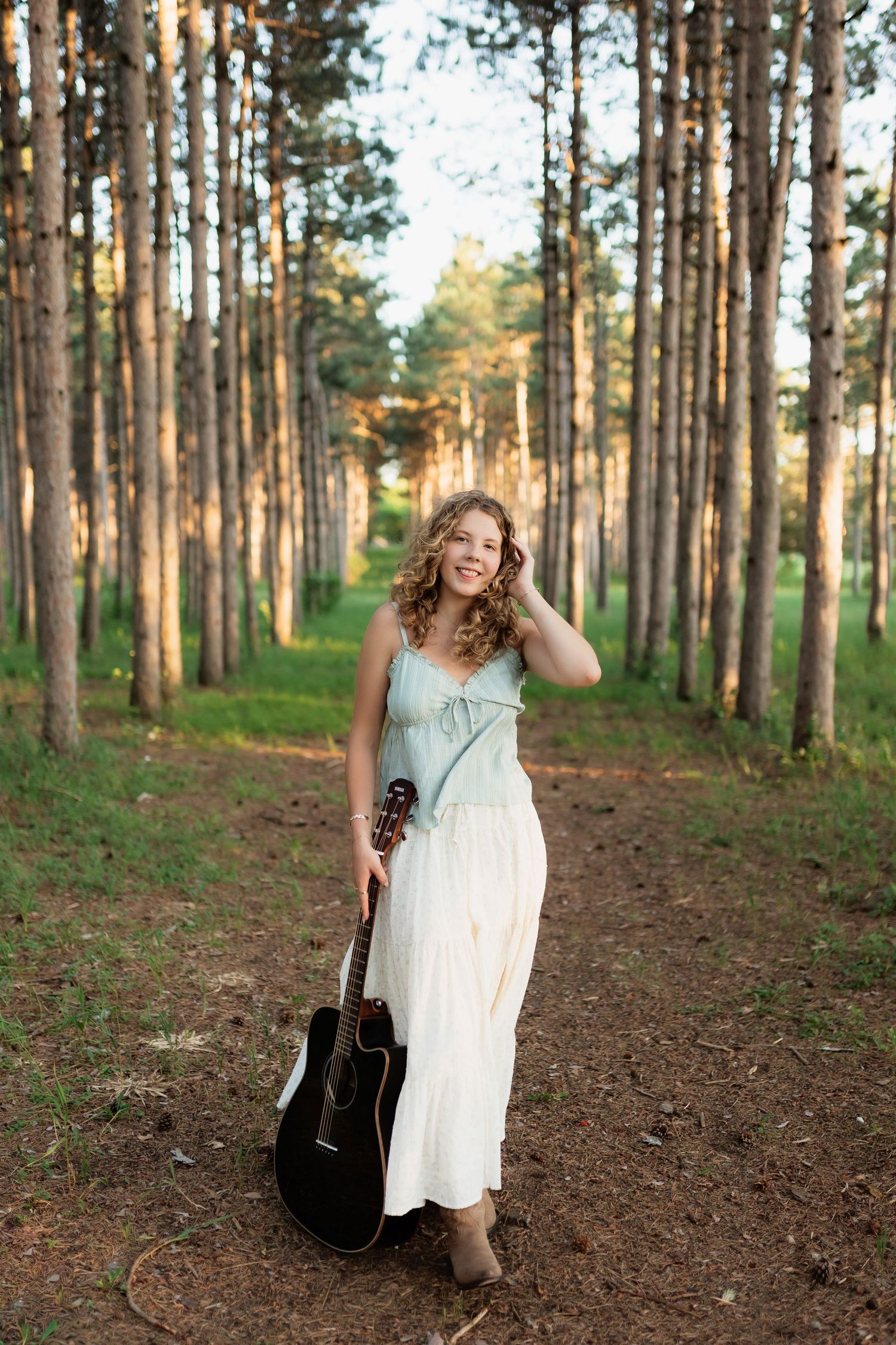 Smiling curly-haired woman holding acoustic guitar while standing in a sunlit pine forest path.
