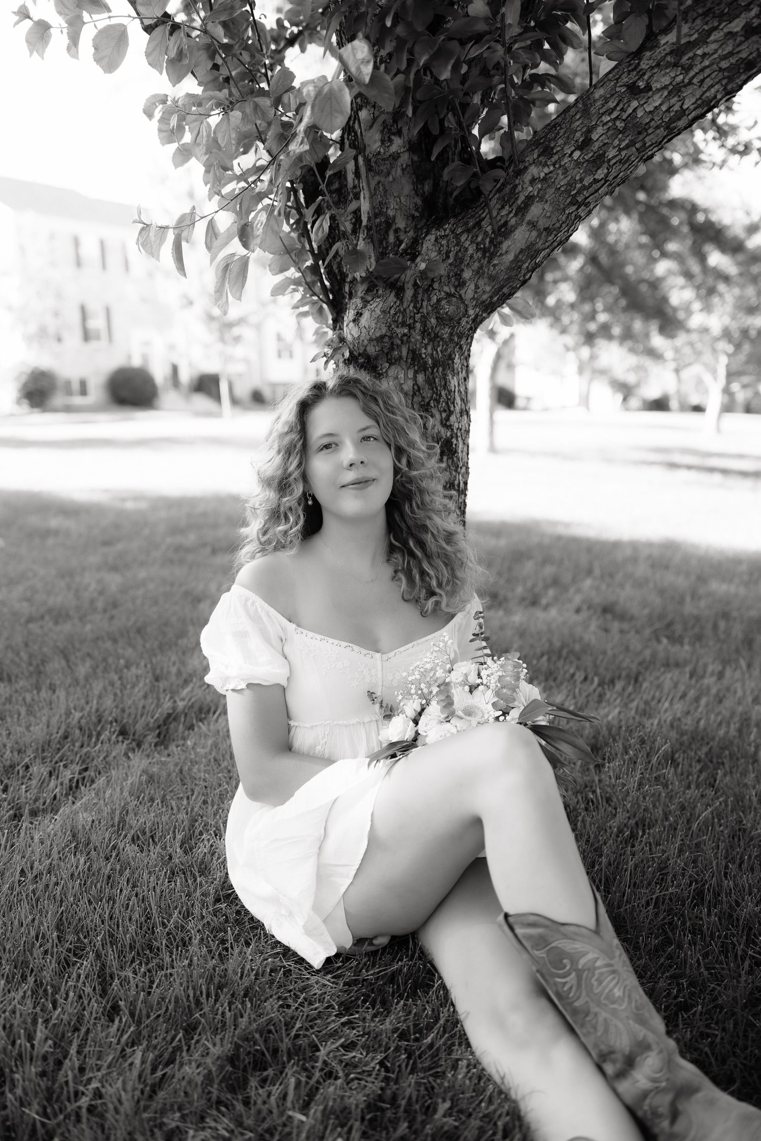 Black and white photo of a young woman in white dress and cowboy boots sitting under a tree holding flowers.