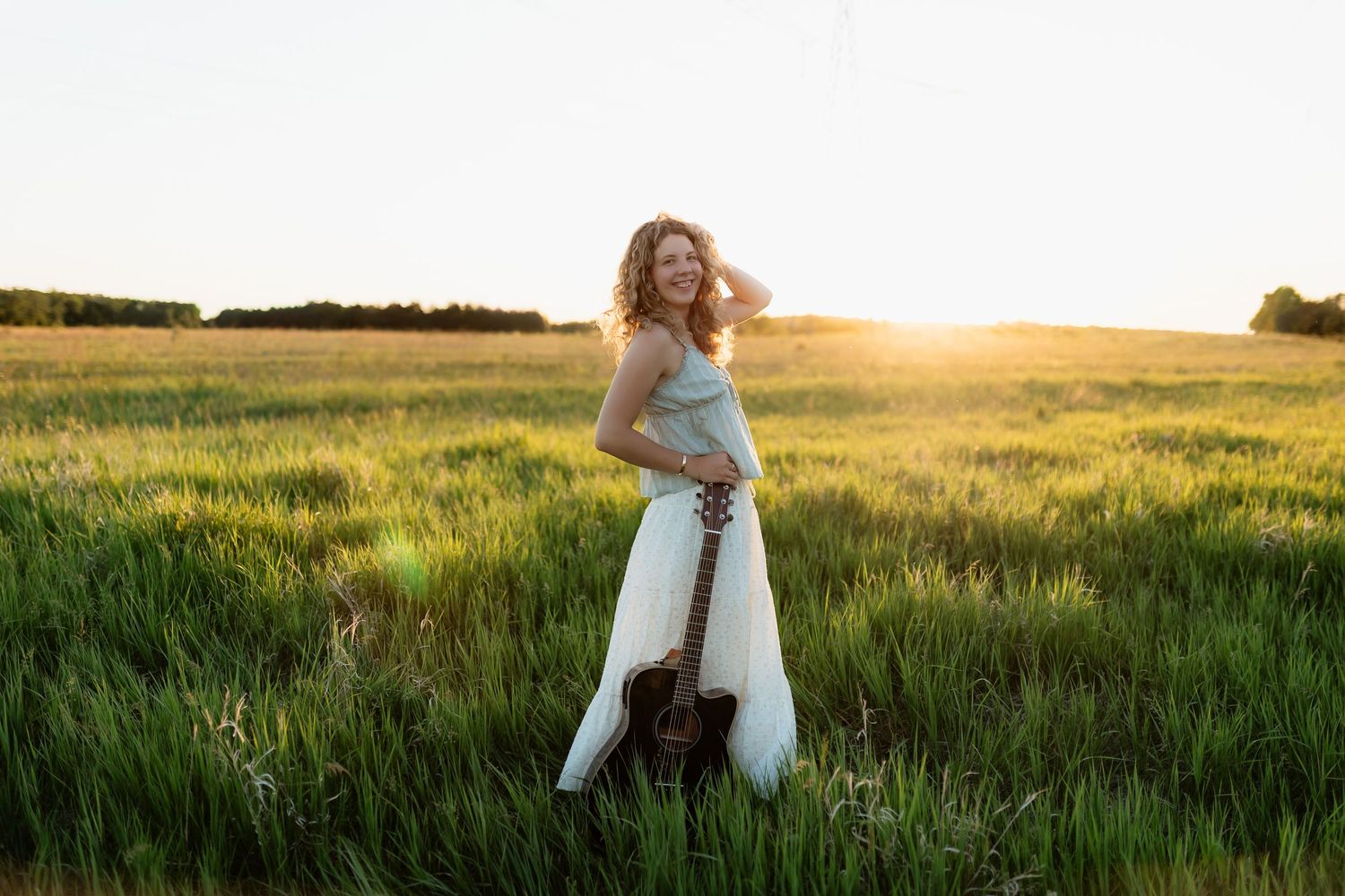 Woman in white dress holding acoustic guitar standing in green field during golden sunset hour.