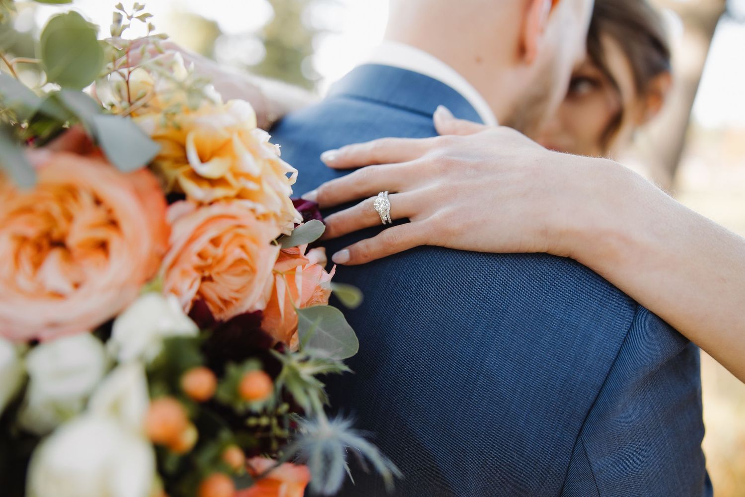 Close-up of bride's hand with diamond ring resting on groom's blue suit jacket with orange roses bouquet.