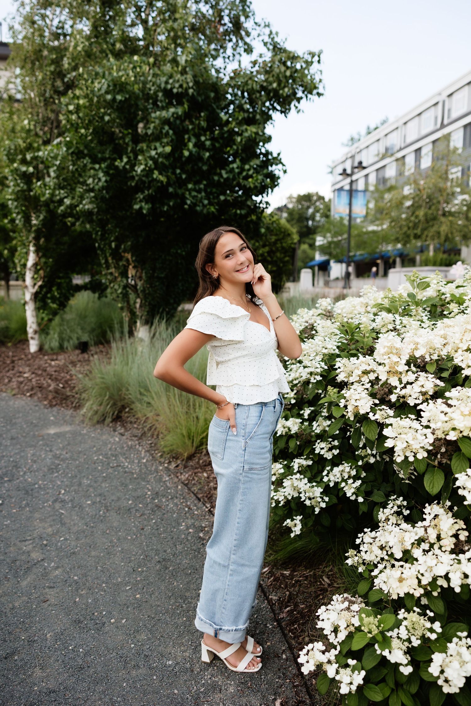Young woman in white top and light jeans poses beside white flowering bushes in an urban garden setting.