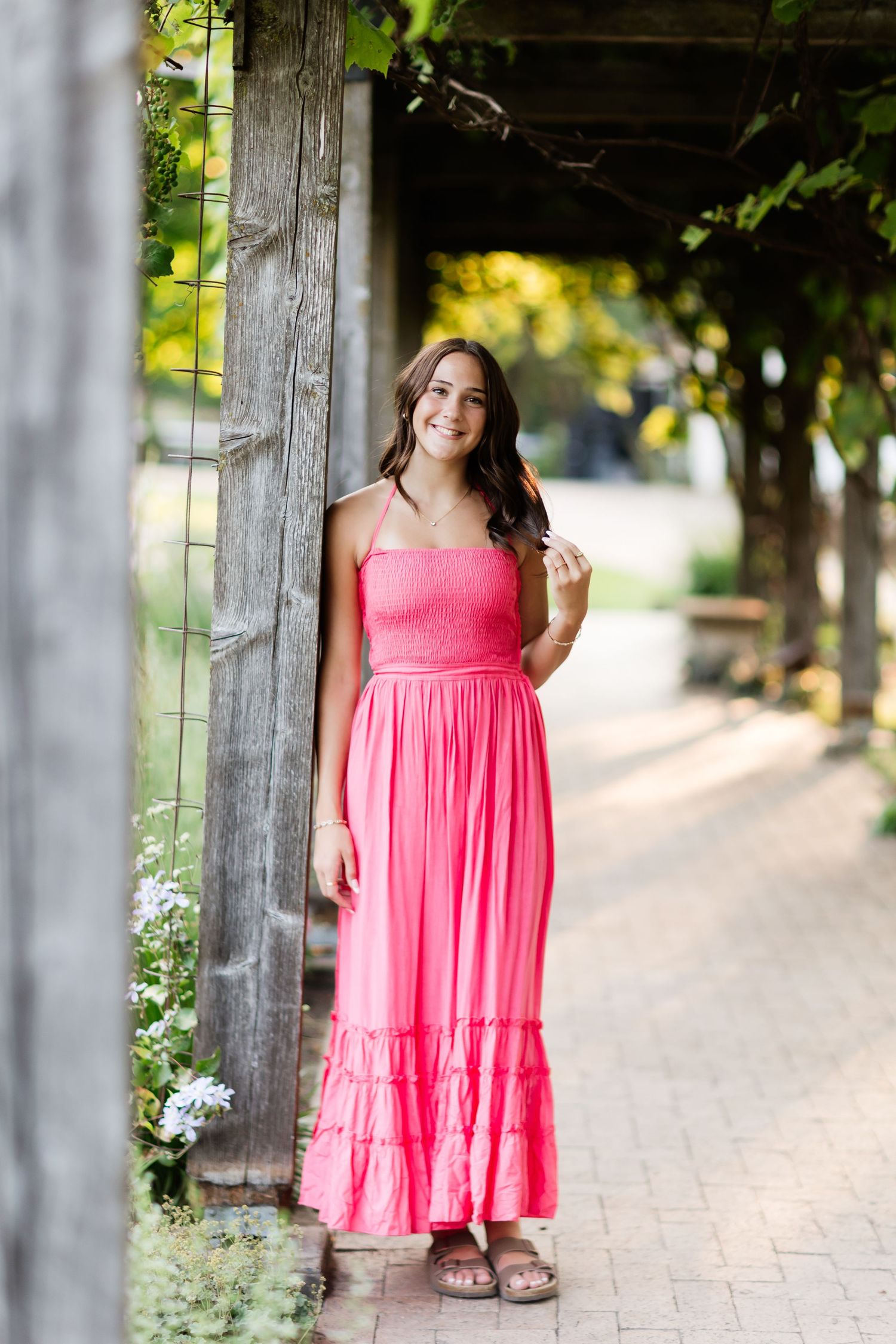 Young woman in a bright pink maxi dress smiling under a rustic wooden pergola surrounded by greenery.