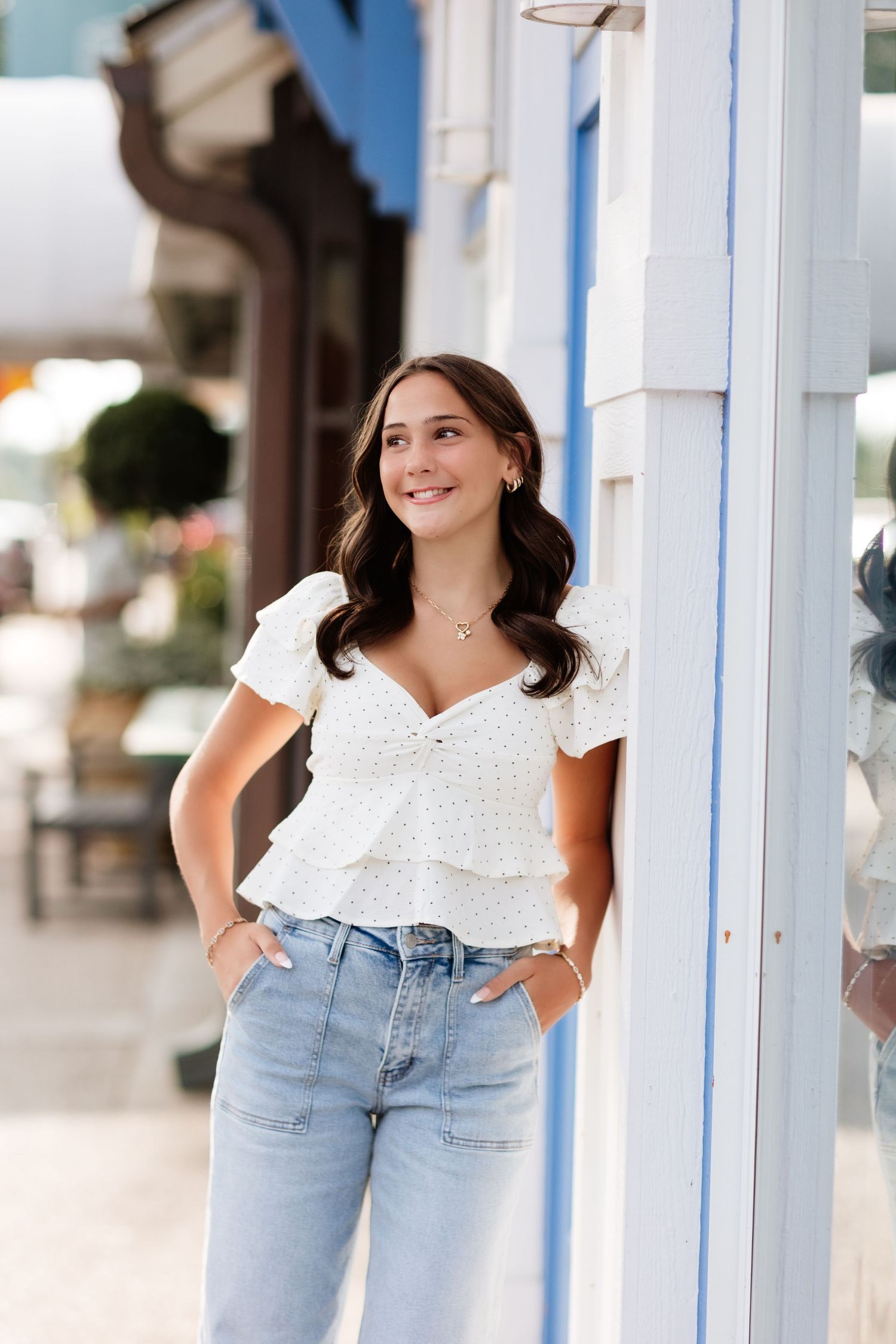 Smiling brunette woman in white peplum top and light jeans leaning against blue and white storefront.