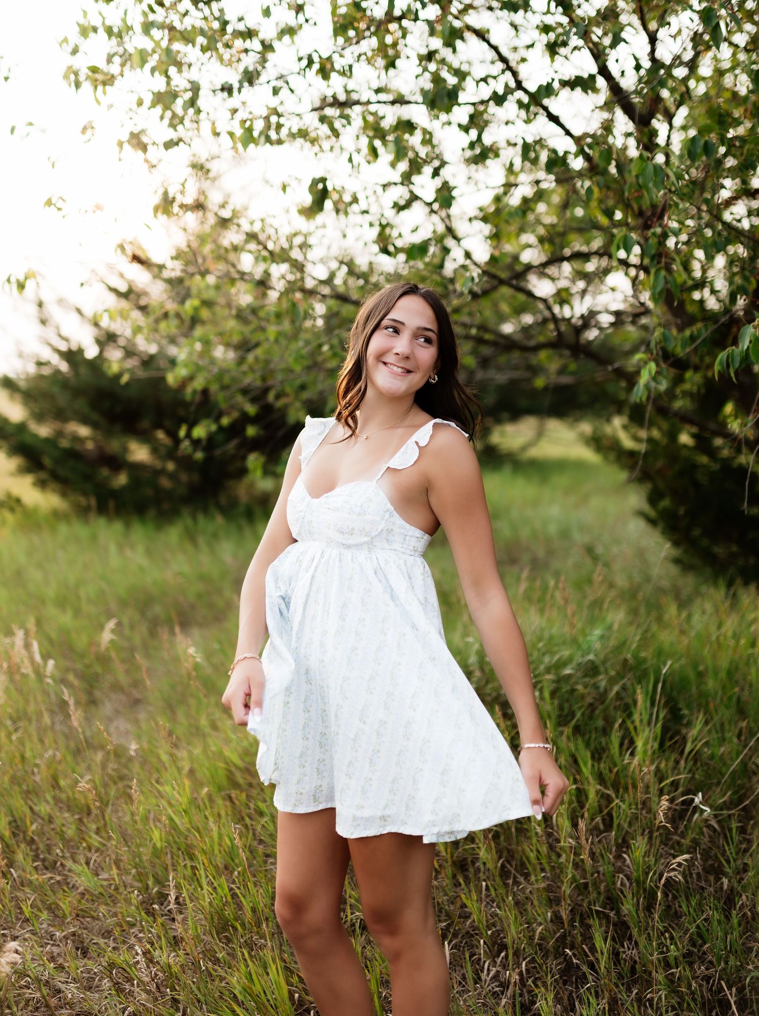 Smiling young woman in white ruffle sundress posing in lush green meadow with flowering trees behind her.