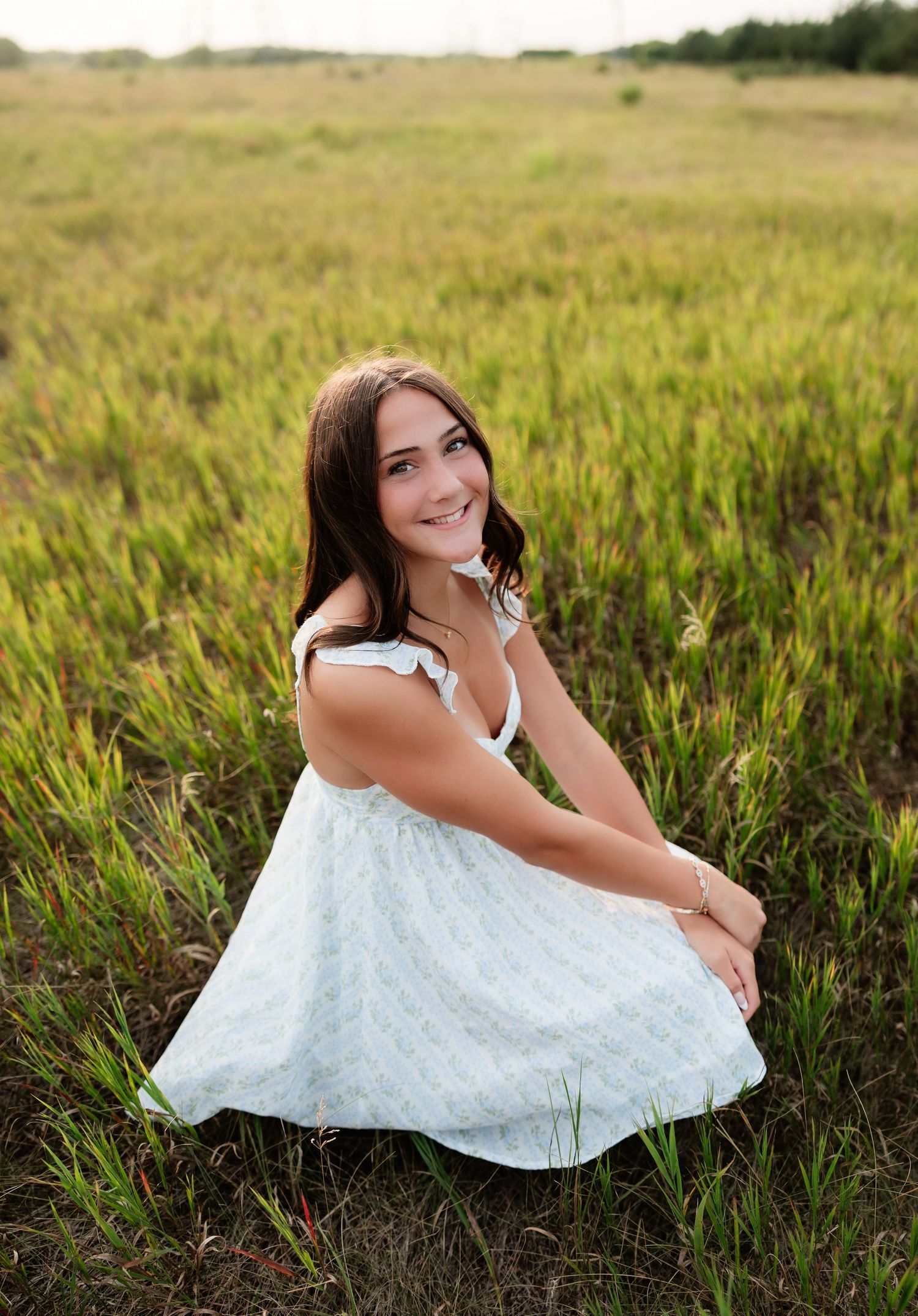 Young woman in white sundress crouching in a green grassy field, smiling over her shoulder.