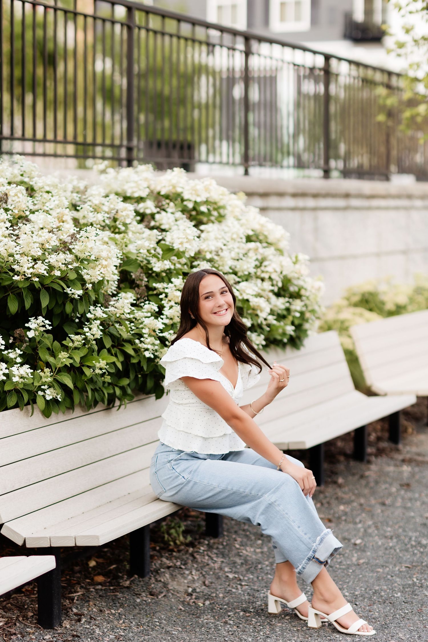 Smiling woman in white ruffled top and jeans sitting on outdoor bench beside white flowering shrubs.