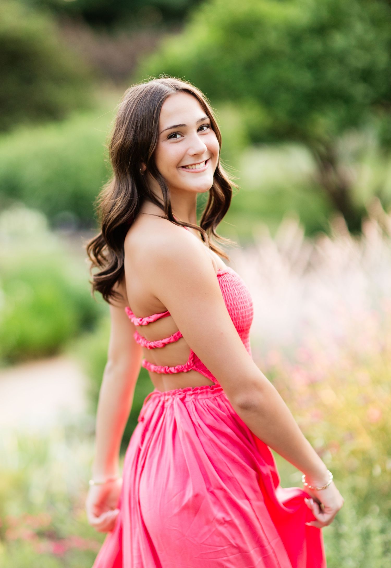 Young woman in coral pink halter dress smiling over shoulder in blooming garden.