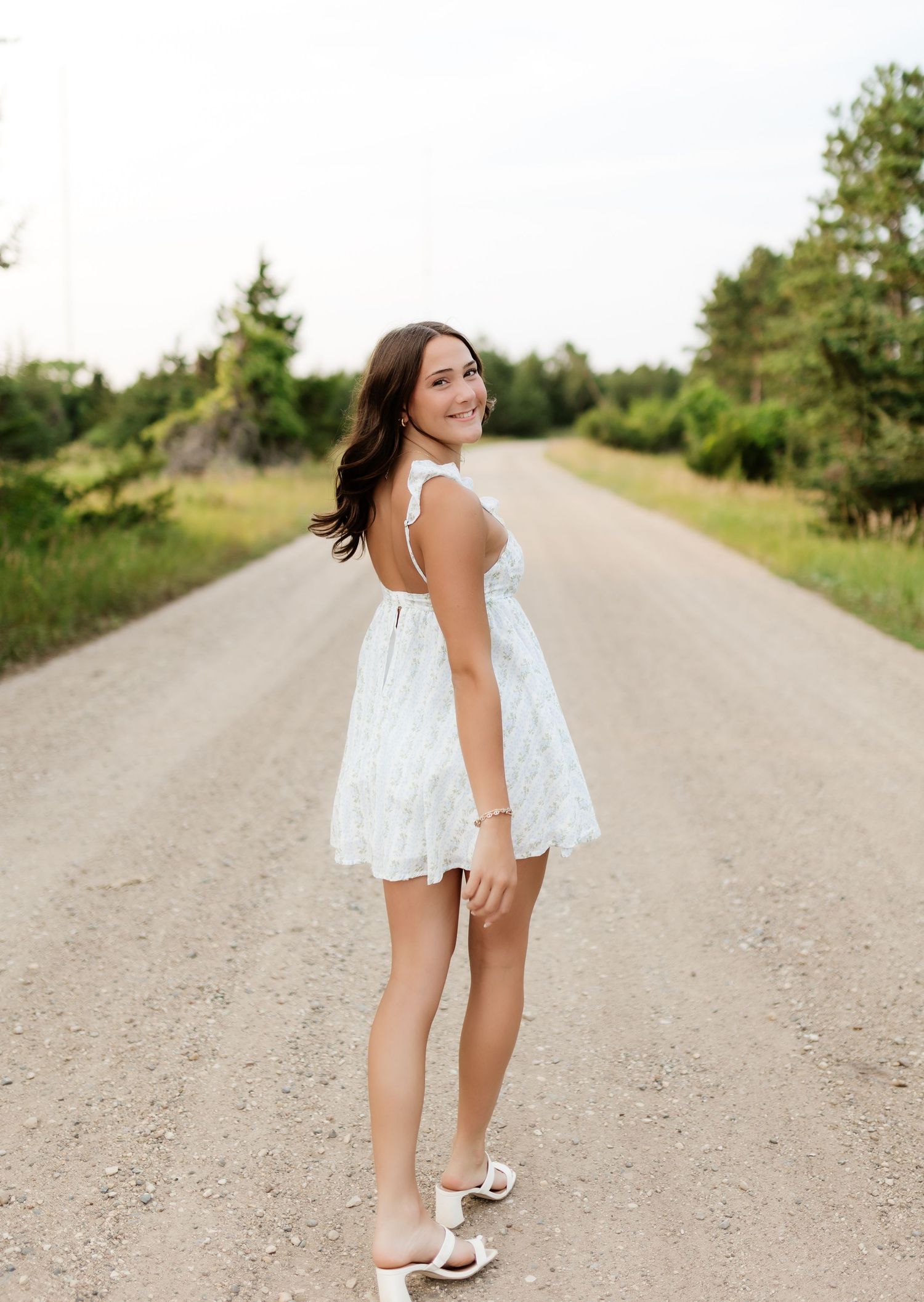 Smiling young woman in white mini dress standing on rural dirt road surrounded by trees.