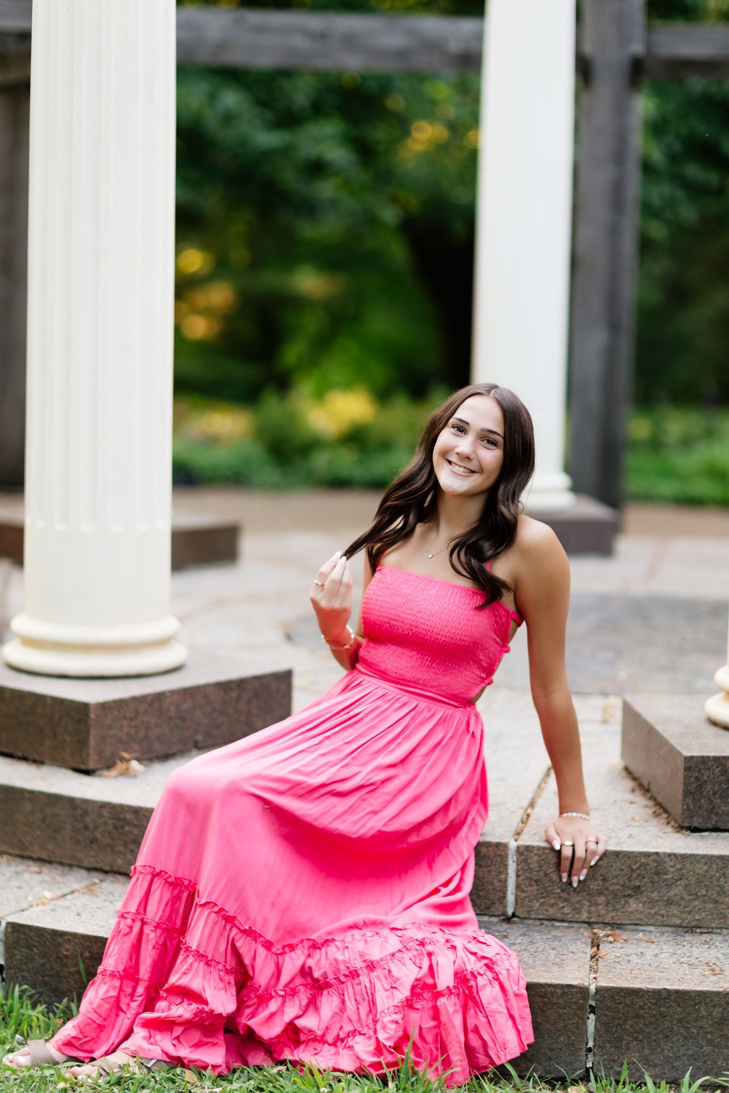 Smiling young woman in pink ruffled maxi dress sitting on stone steps near white columns outdoors.