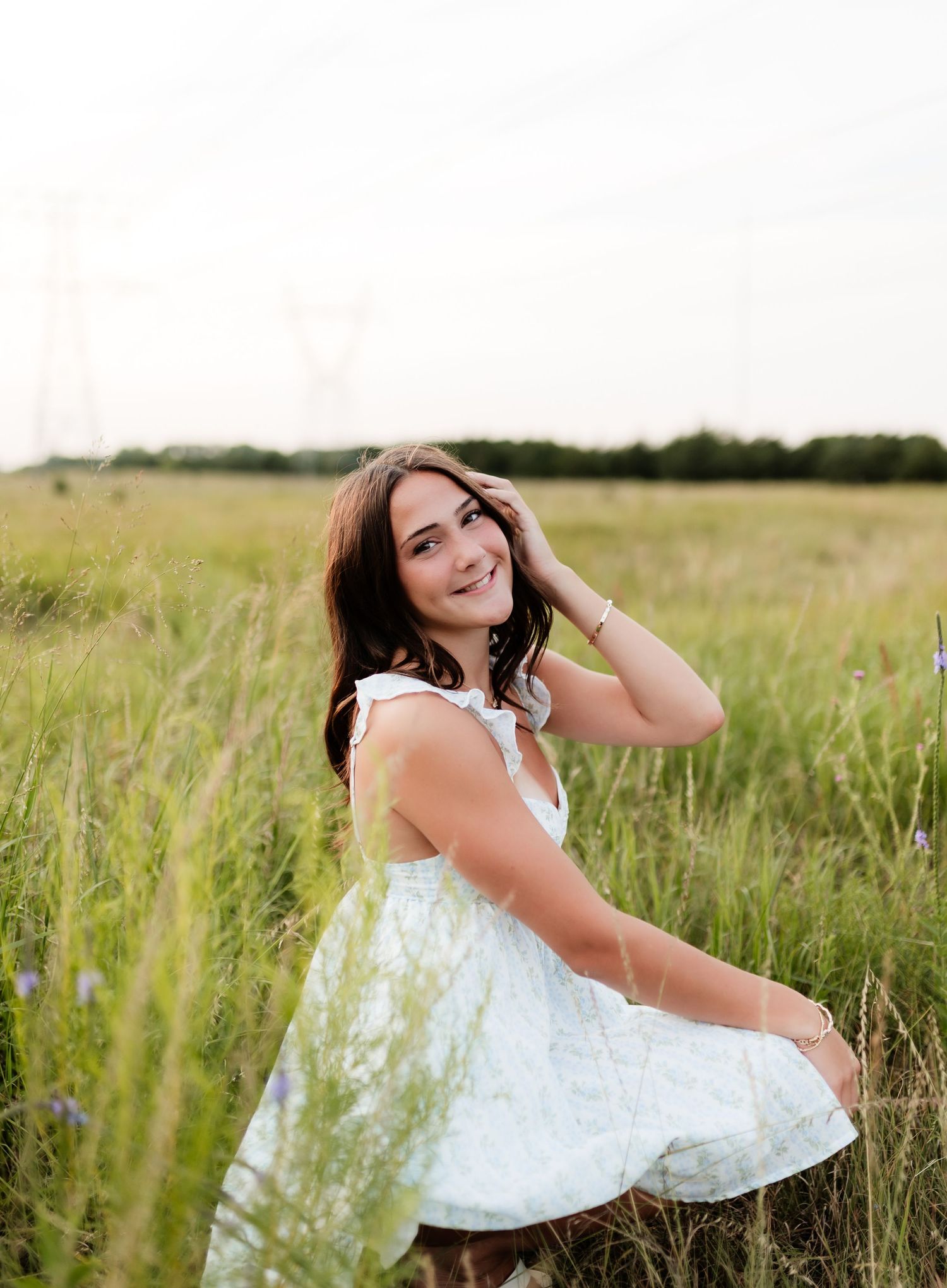 Young woman in white sundress sitting in tall grass field, smiling with hand in hair.
