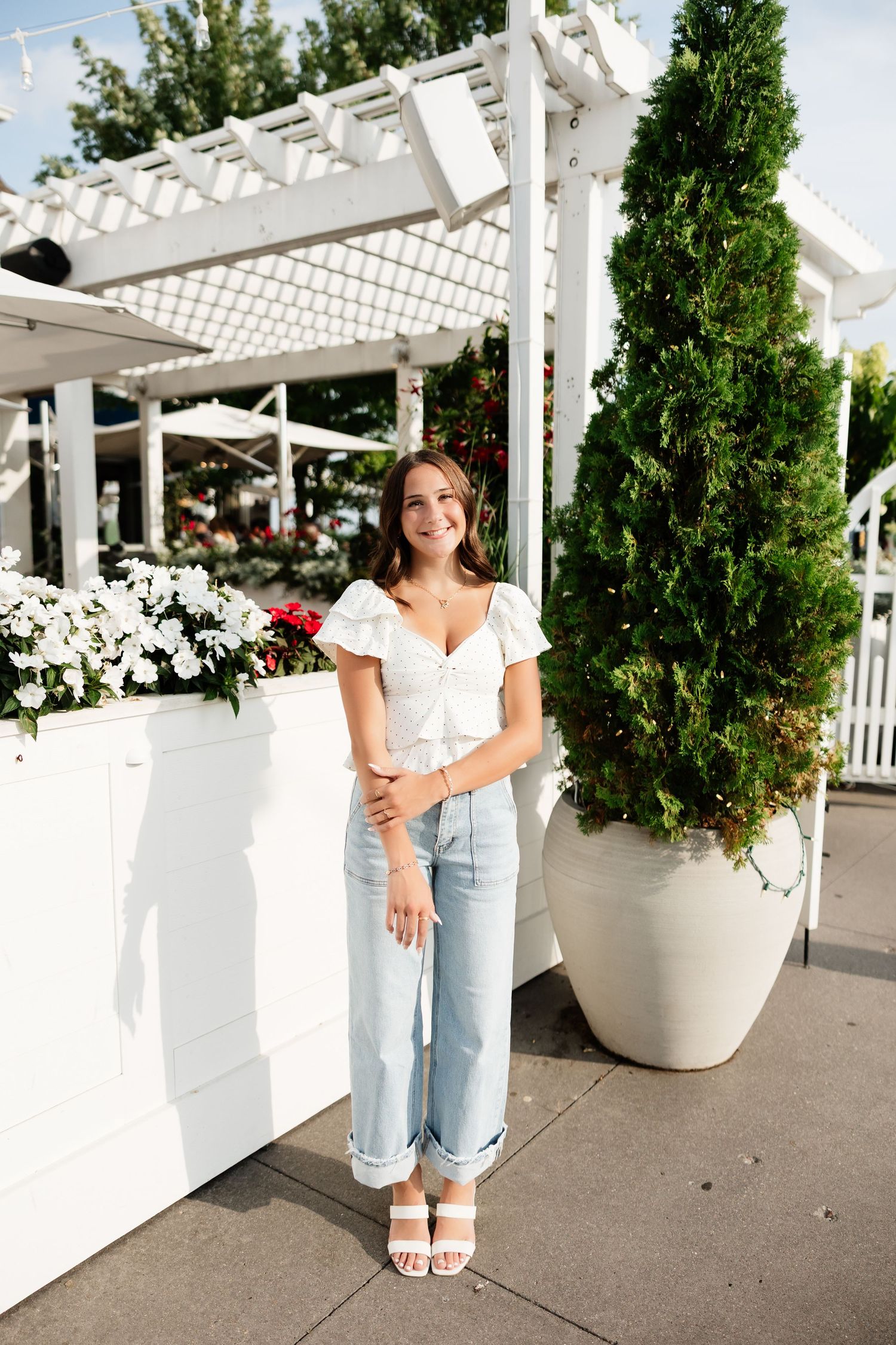 Young woman in white crop top and jeans smiling outdoors near white pergola and greenery.