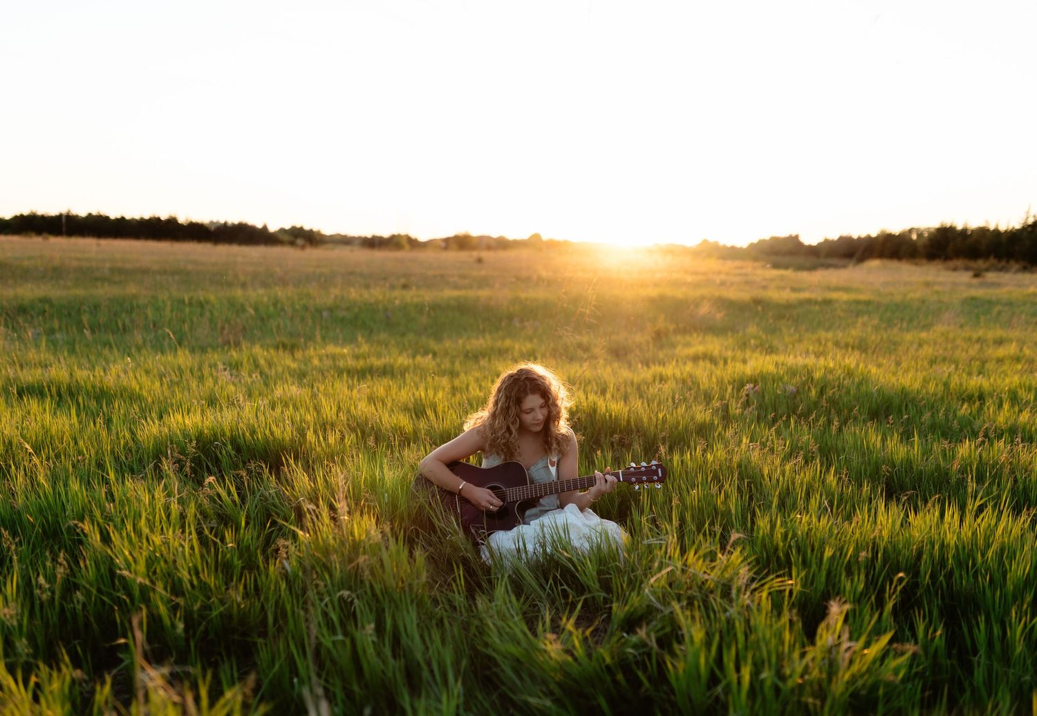 Young woman in white dress playing acoustic guitar while sitting in green field during golden sunset.