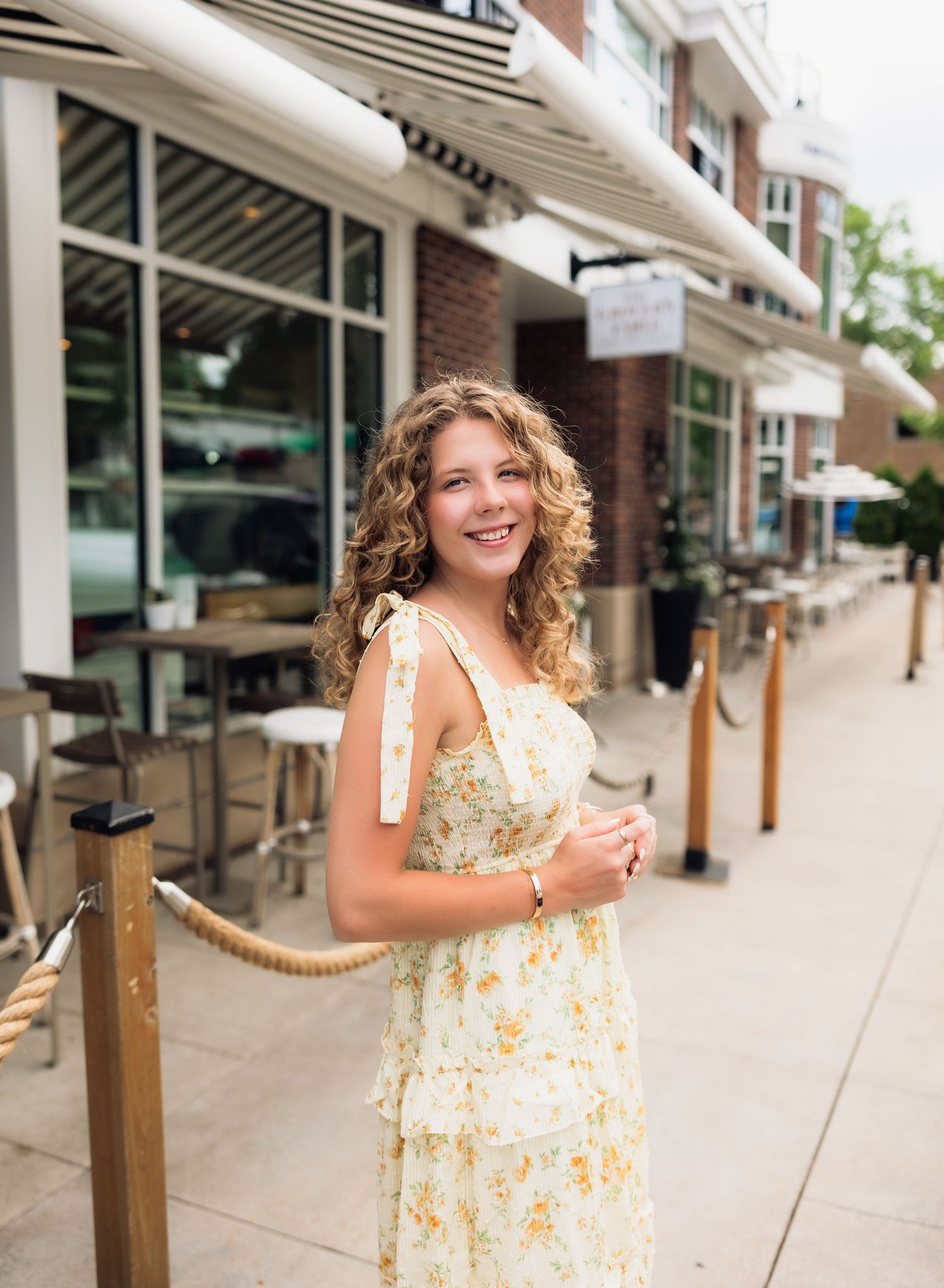 Young woman with curly hair smiling in a yellow floral sundress on a sunny outdoor shopping street.