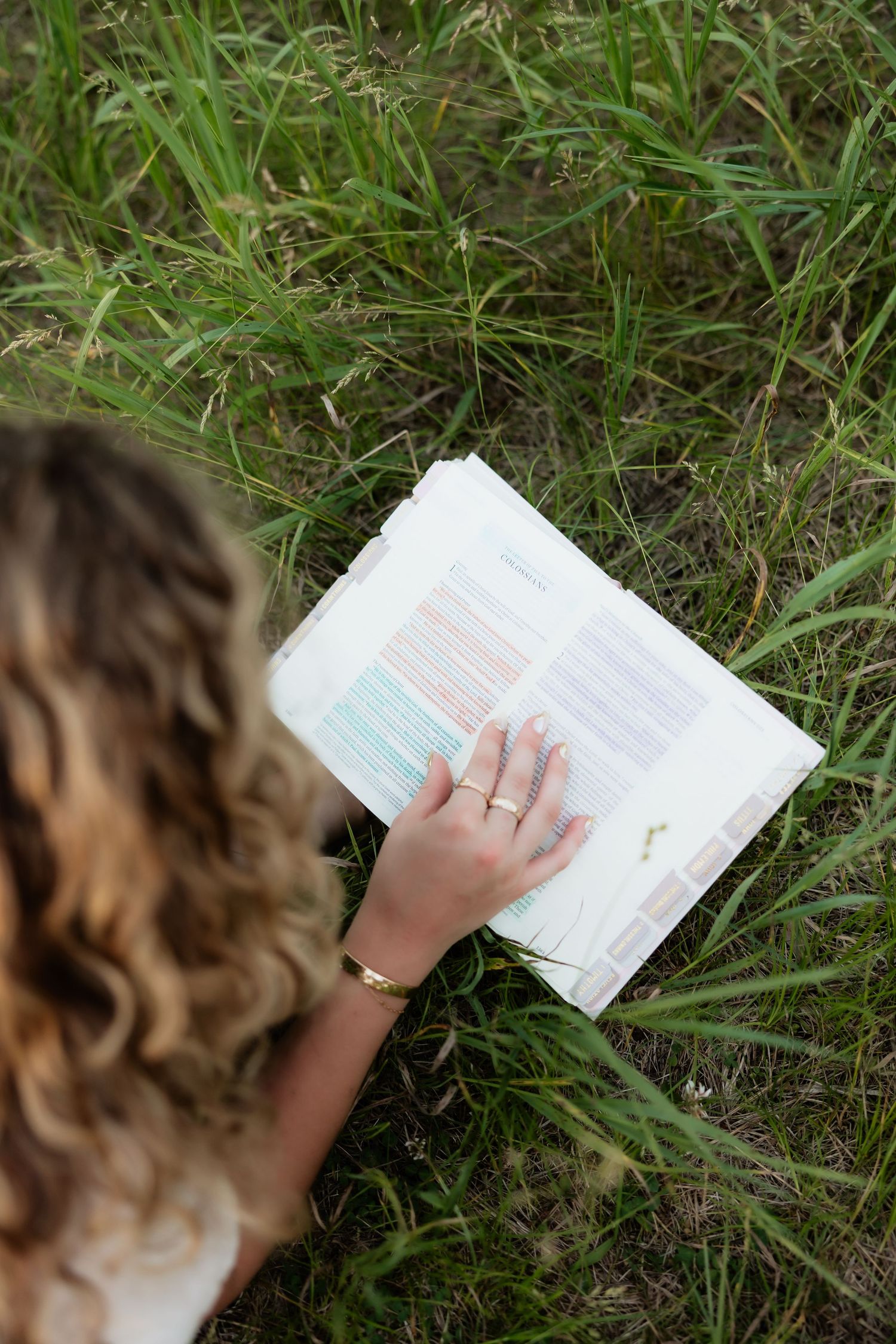 Overhead view of a woman with curly hair reading a colorful Bible in green grass.