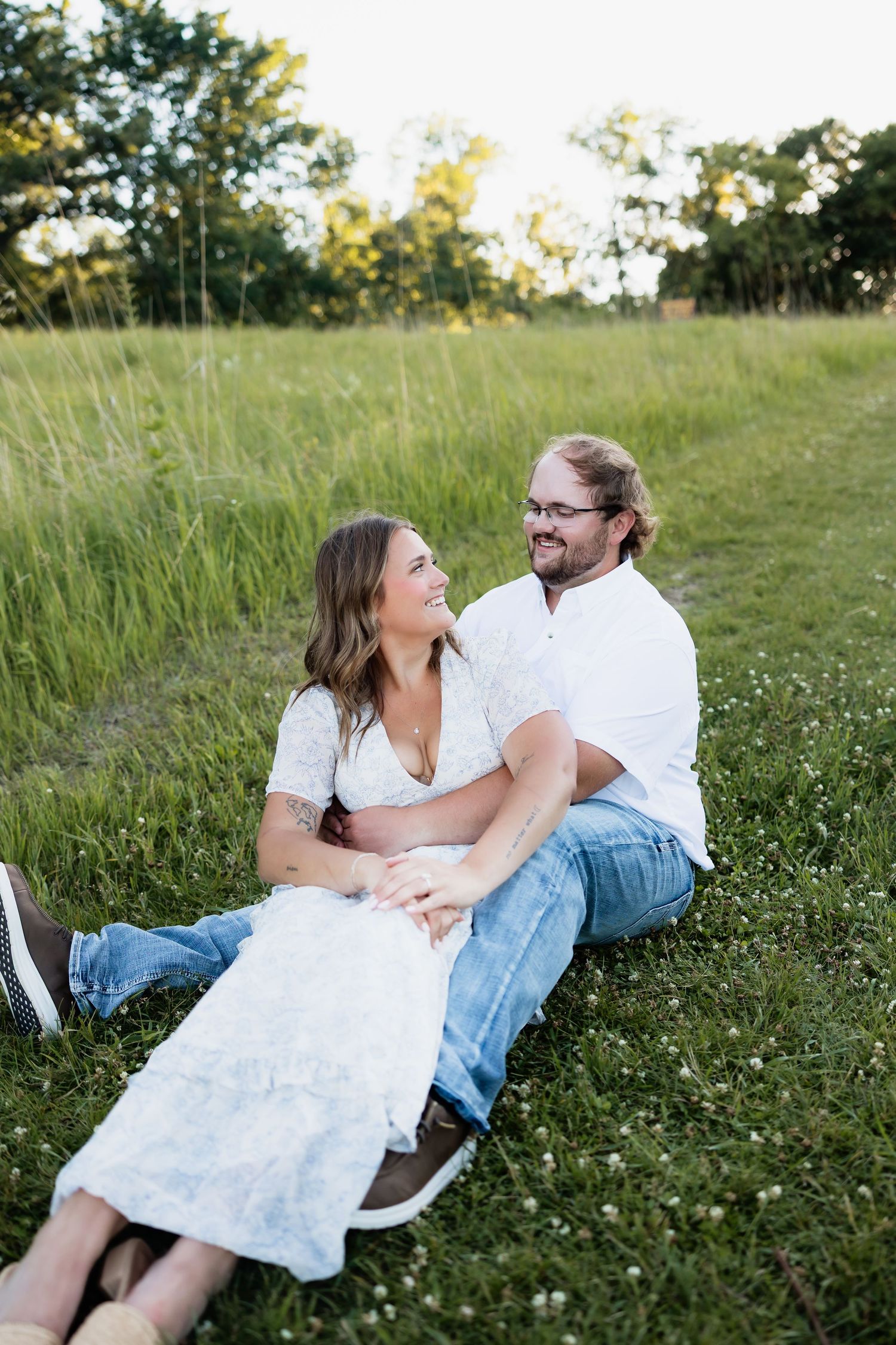 A couple sits together in a lush green meadow, smiling and laughing, the woman in a white dress and the man in jeans.