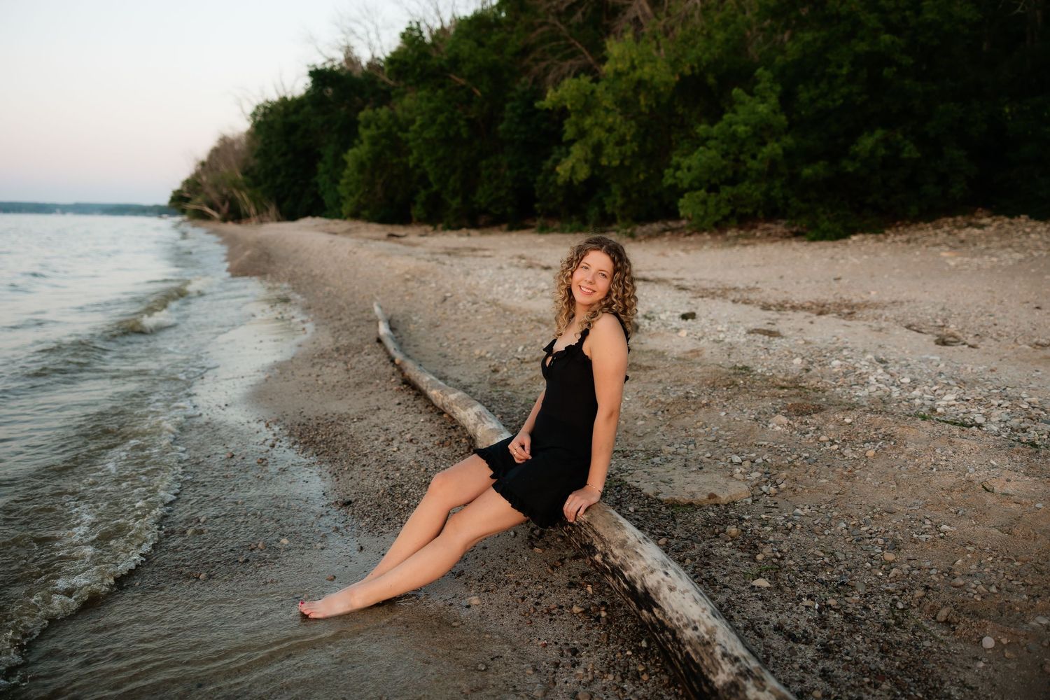 Young woman in black dress sitting on driftwood log on sandy beach at dusk with trees and calm water behind her.