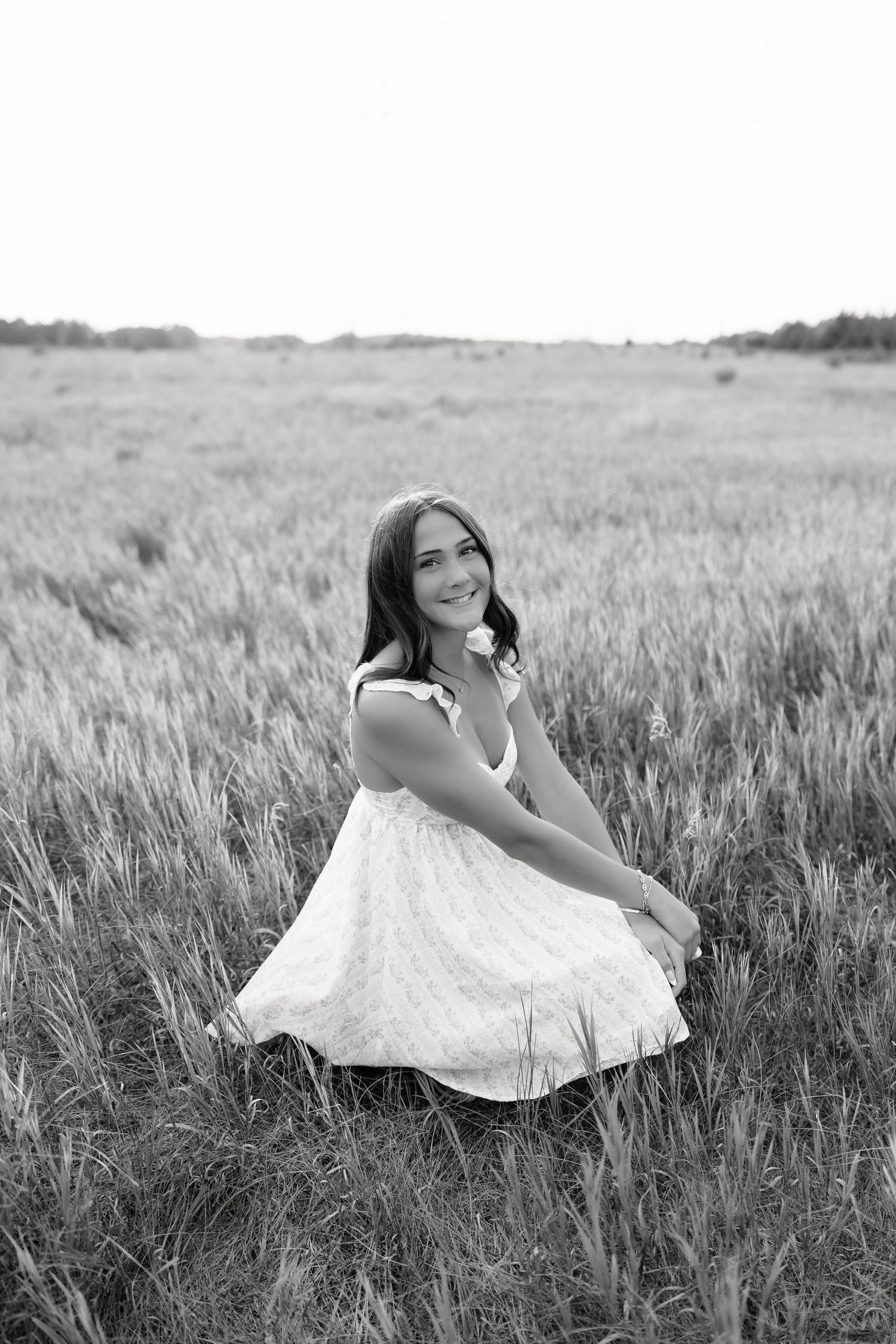 Black and white portrait of a smiling young woman in a floral dress crouching in a wide open grassy field.