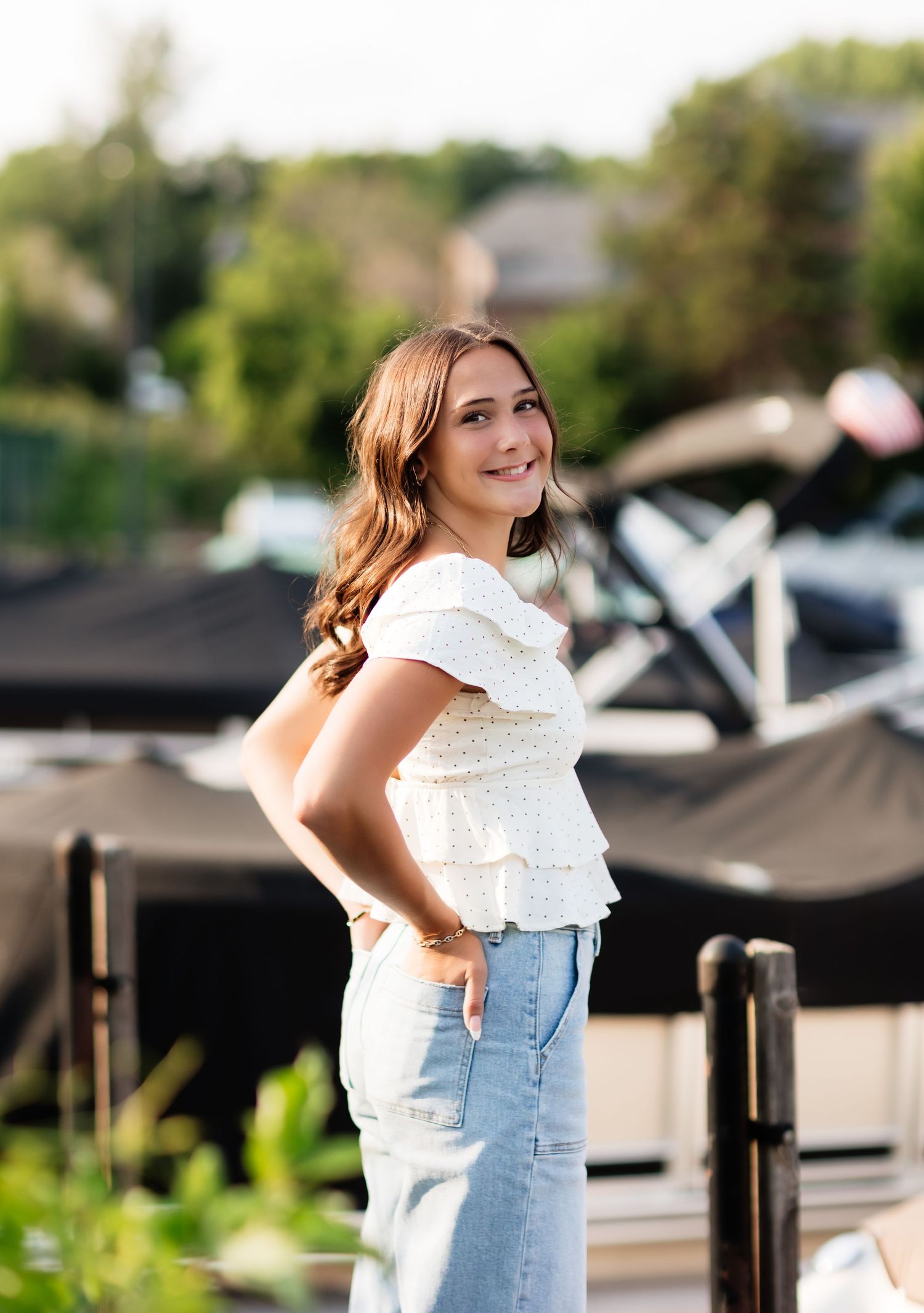 Smiling young woman in white ruffled top and light denim jeans posing near marina with boats.