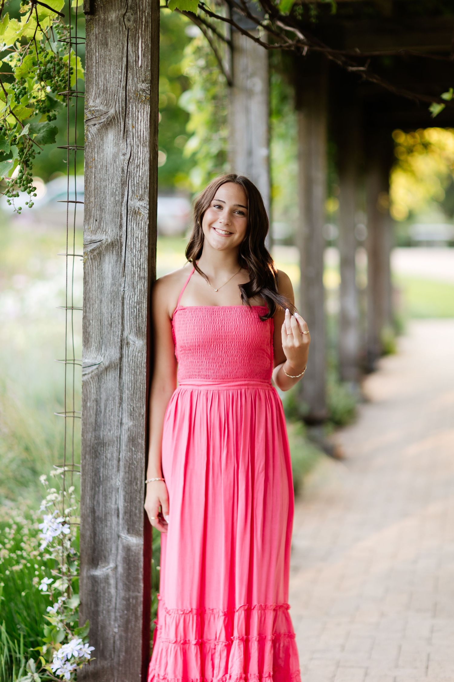 Young woman in coral pink maxi dress leaning against a rustic wooden pergola post in a garden.