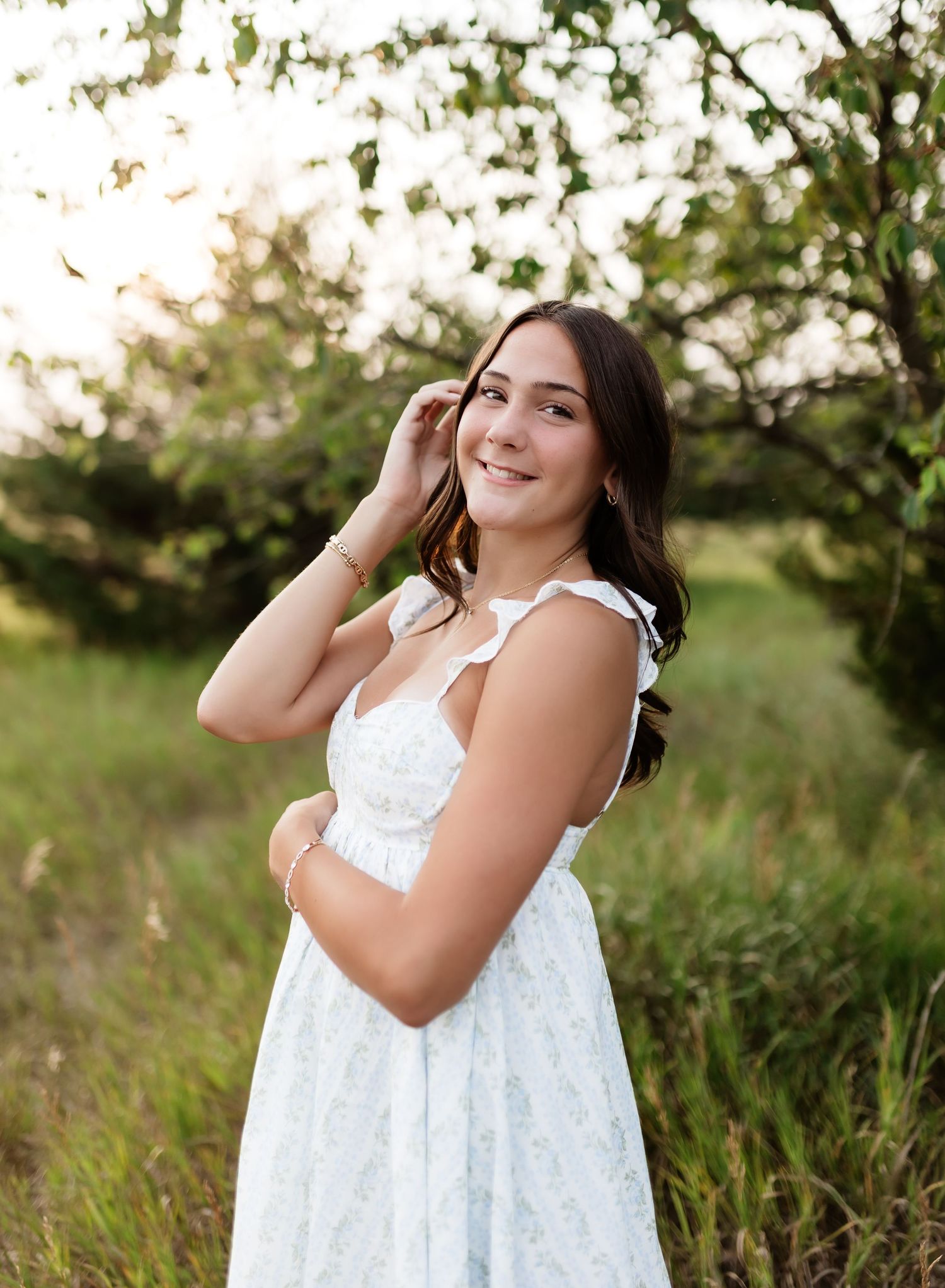 Smiling young woman in floral white dress posing in green outdoor setting.