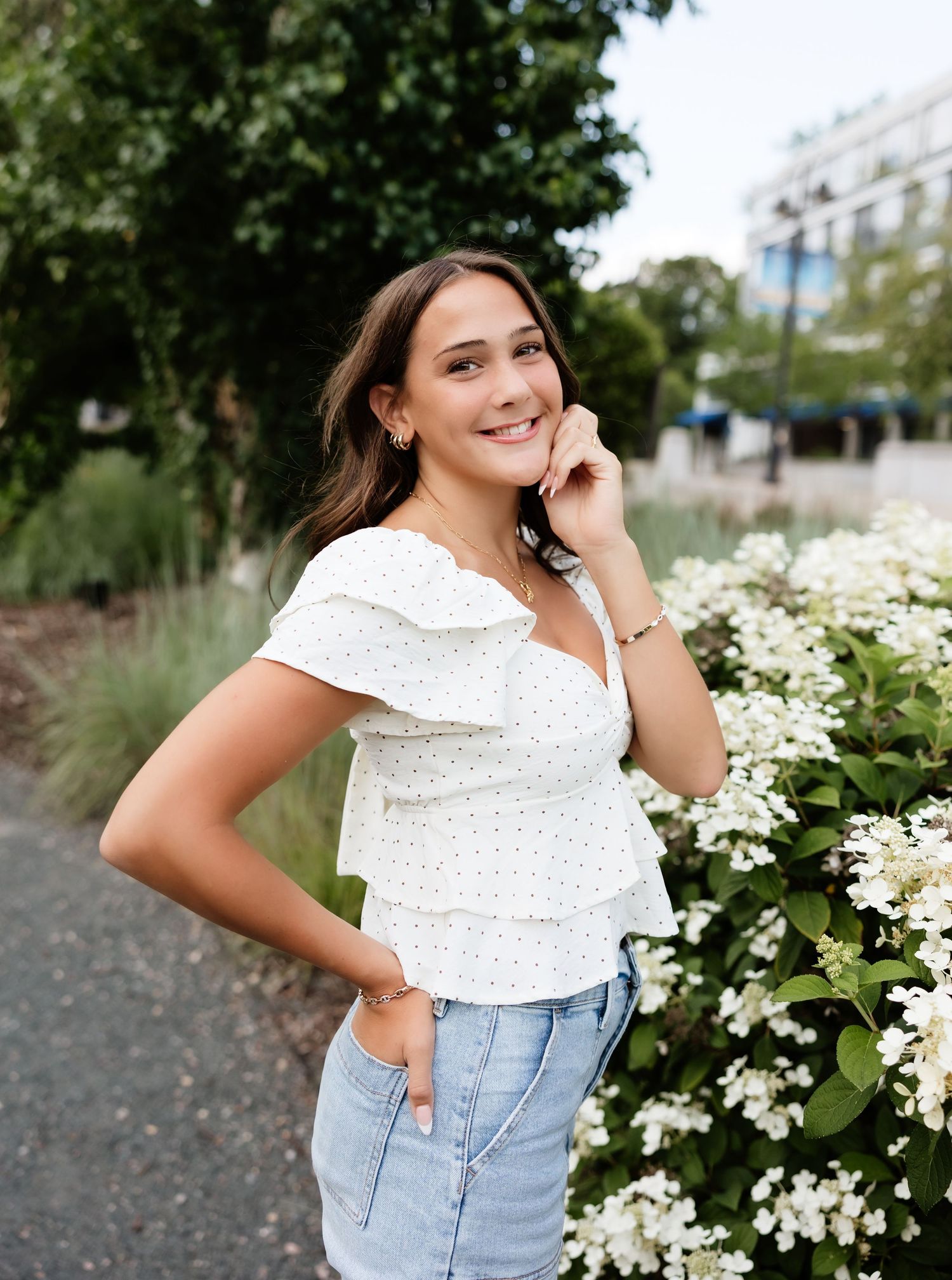 Young woman in white ruffled top and jeans smiling beside white hydrangeas in an outdoor garden setting.
