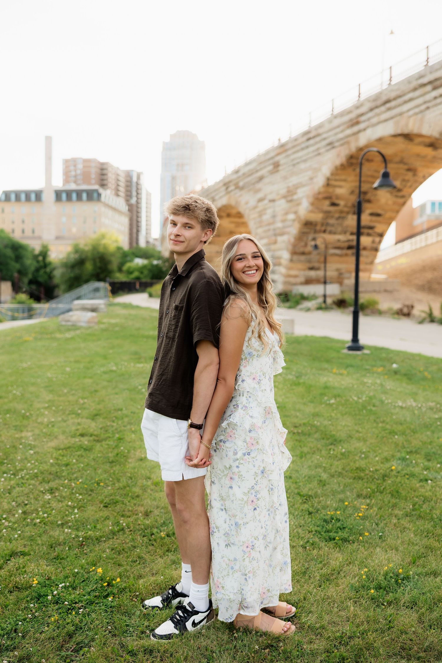A young couple holds hands smiling on green grass near a stone arch bridge and city skyline.