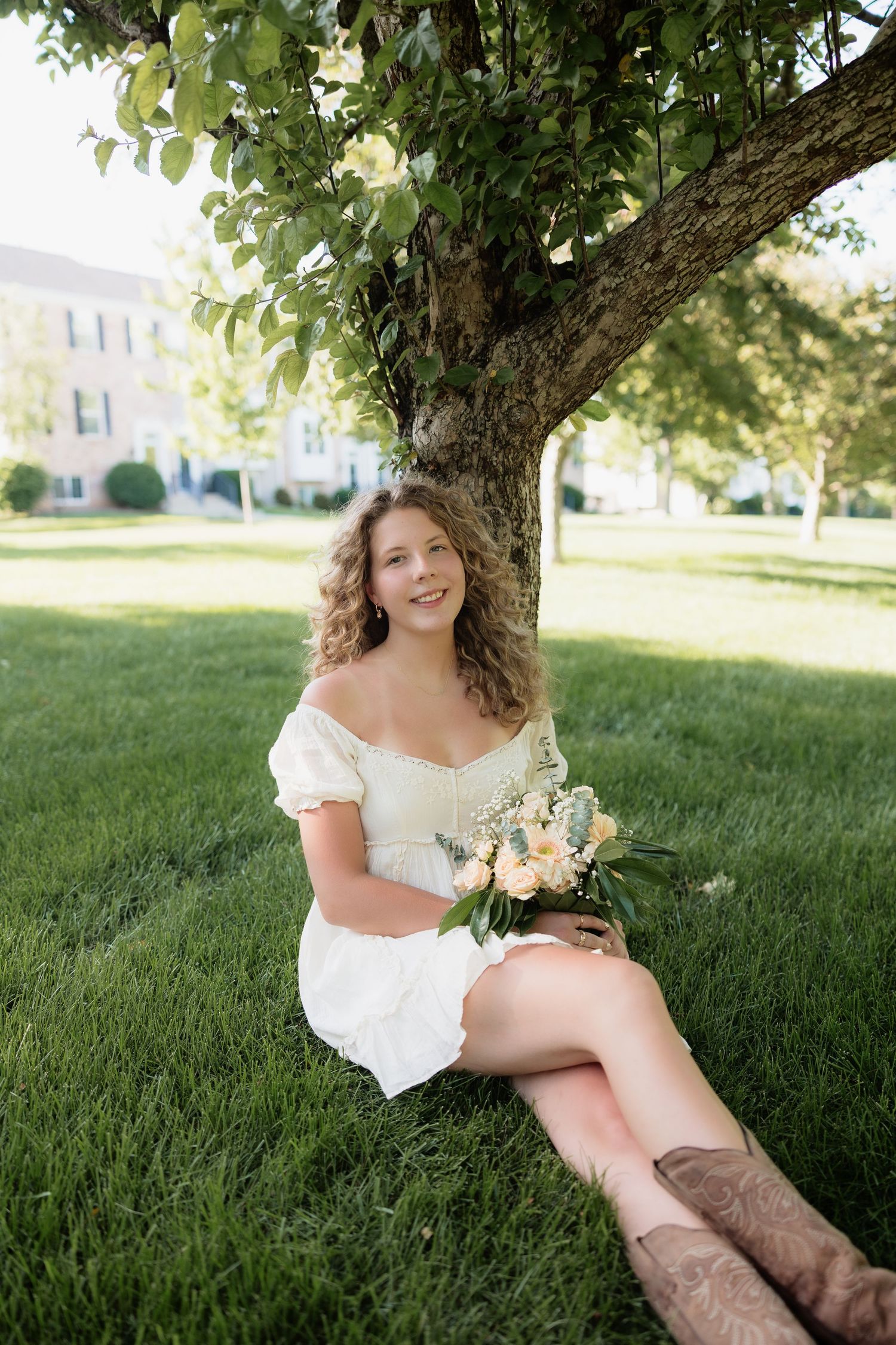 Young woman in white dress sitting under tree holding bouquet of flowers on green grass.