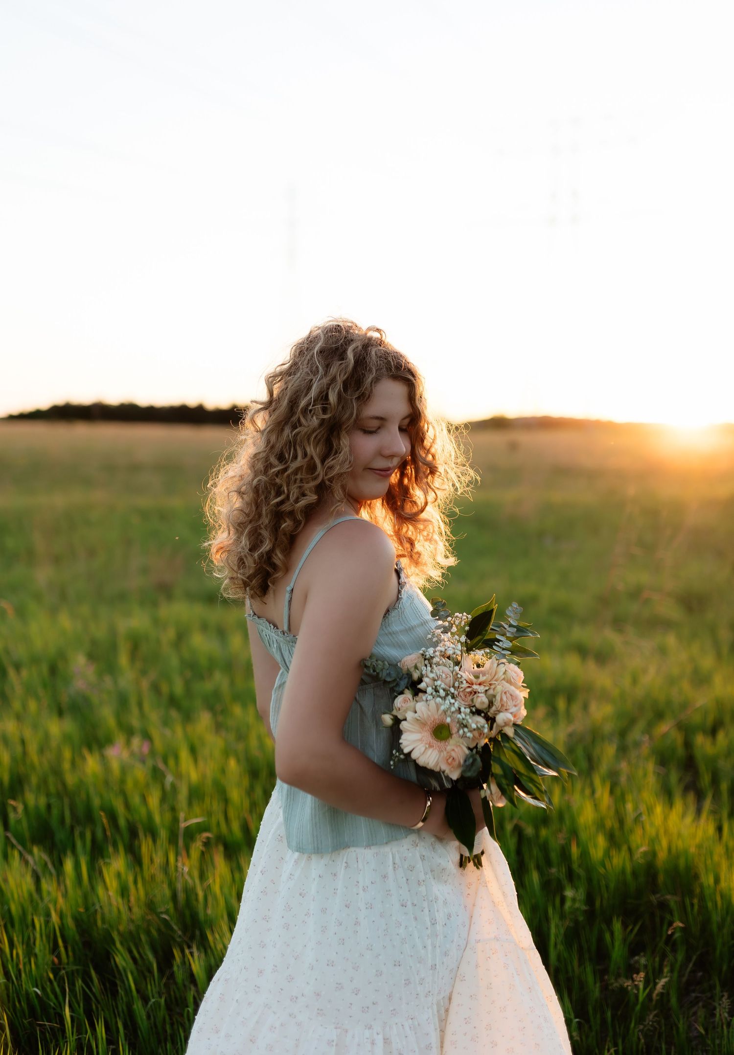 Young woman with curly hair holding a bouquet of white flowers in a green field at golden hour sunset.