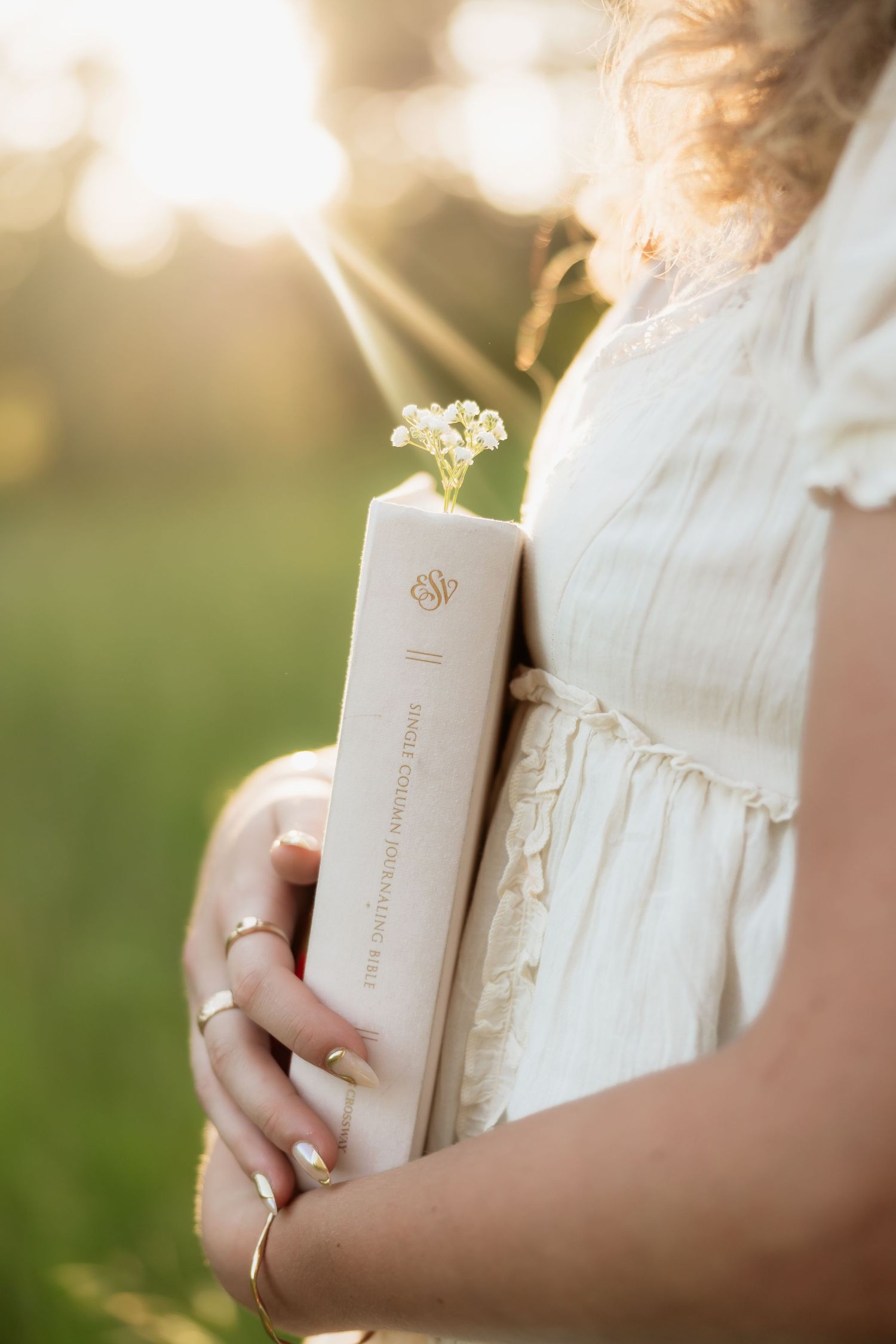 Woman in white dress holding an ESV Bible with small white wildflowers in golden sunset light.