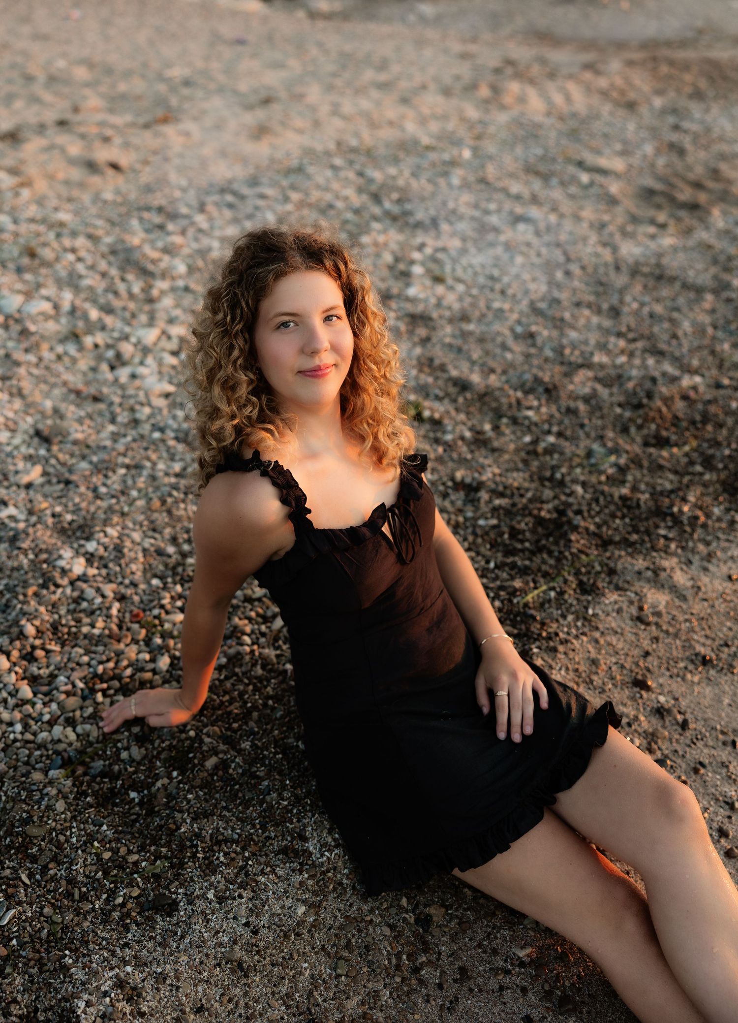 A young woman with curly hair sits on a pebble beach in a black dress during warm golden hour light.