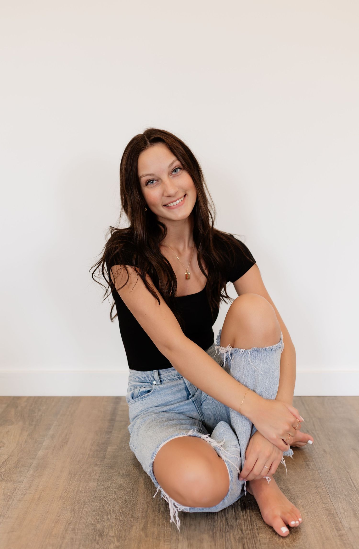 Smiling brunette woman sitting on floor in black tee and ripped denim shorts against white wall.