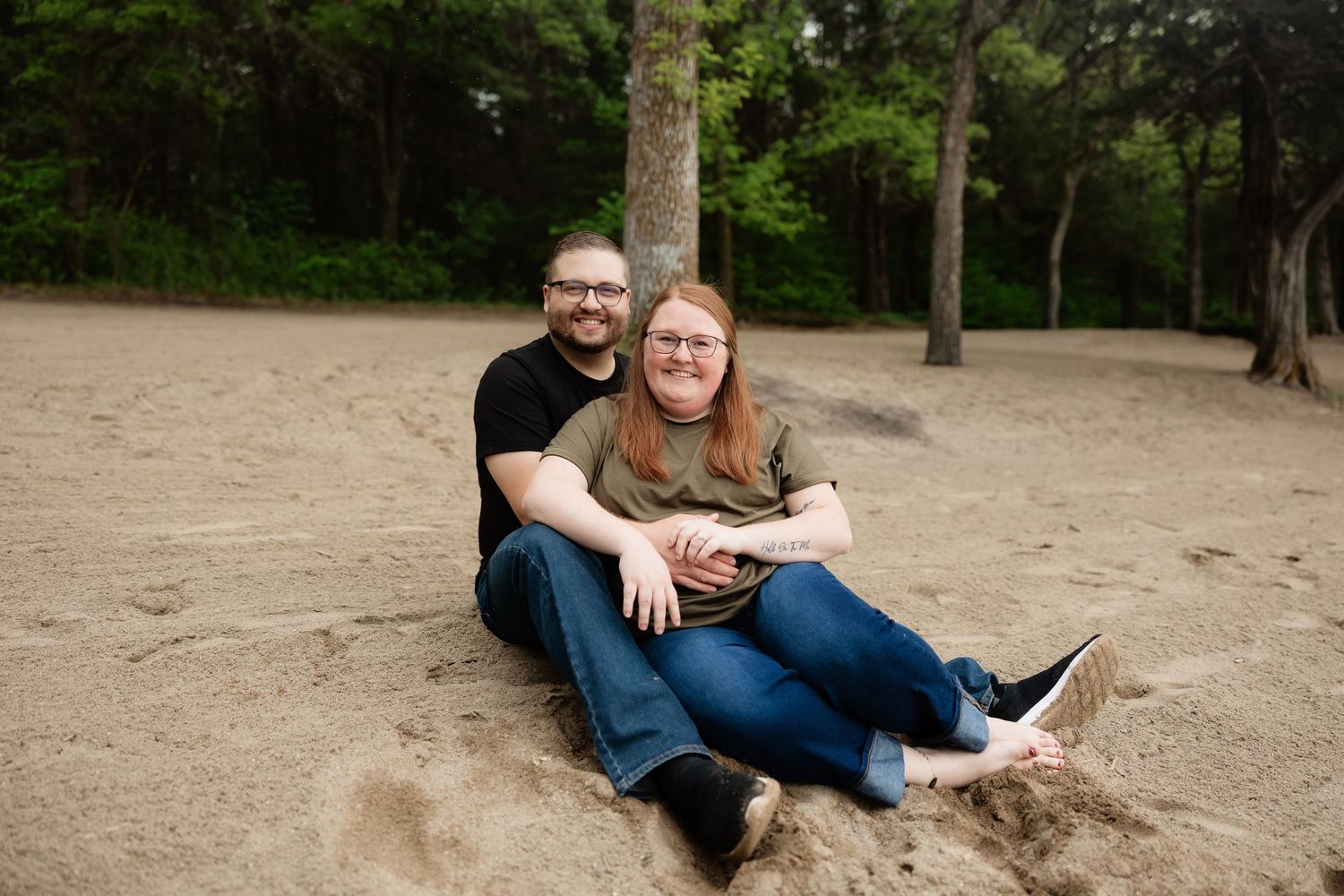 Happy couple sitting together on a sandy beach surrounded by trees, smiling at the camera.