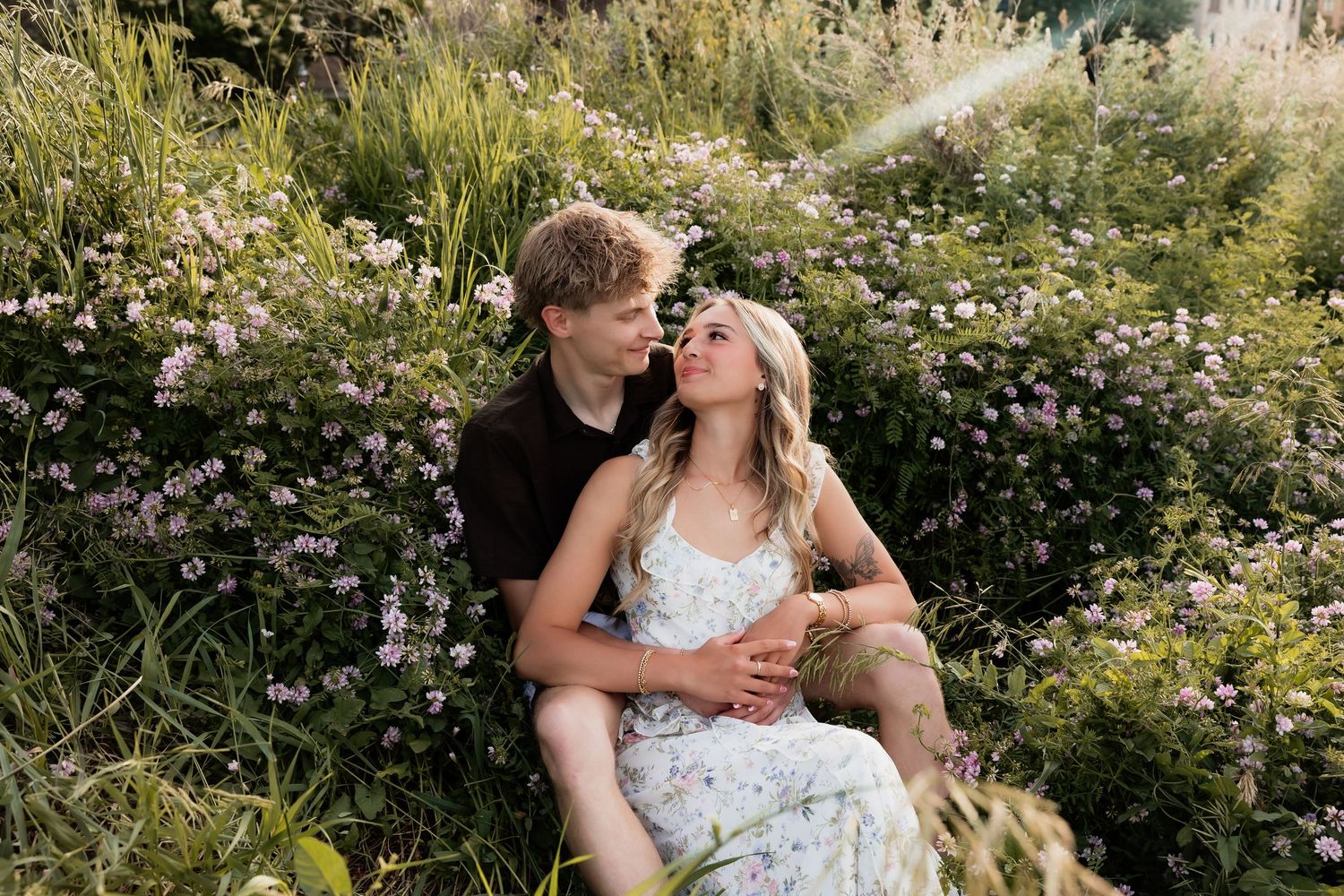 Young couple sitting romantically among wildflowers and lush greenery, sharing a tender moment outdoors.