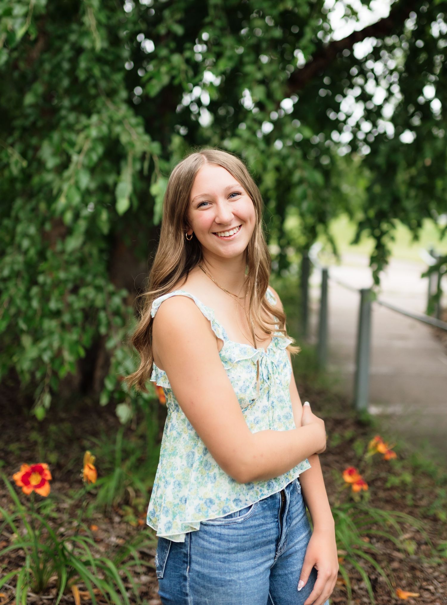 Smiling teenage girl in floral top and jeans posing outdoors near lush green trees and orange flowers.