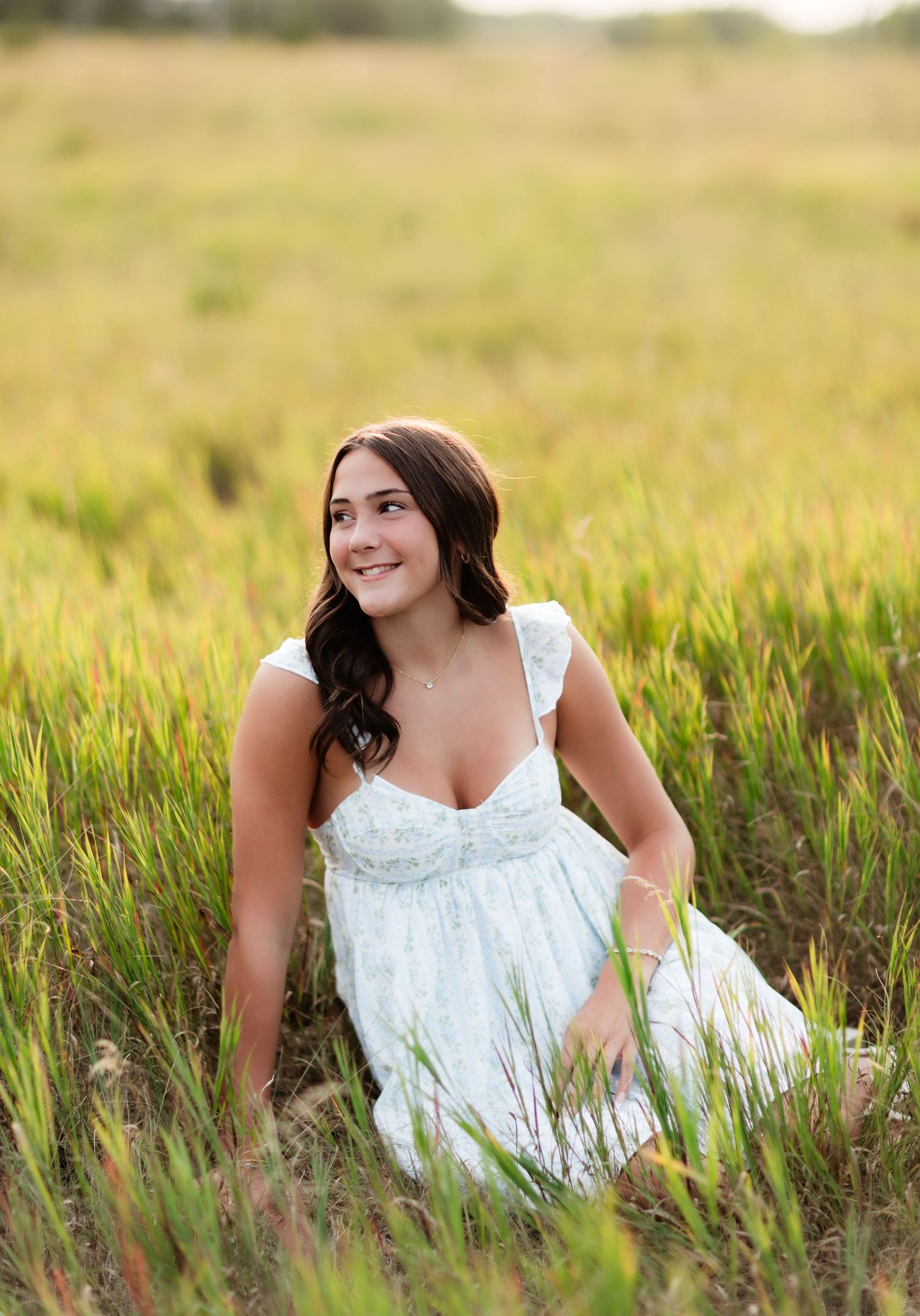 Smiling young woman in a white floral dress sits in a lush green meadow bathed in warm sunlight.