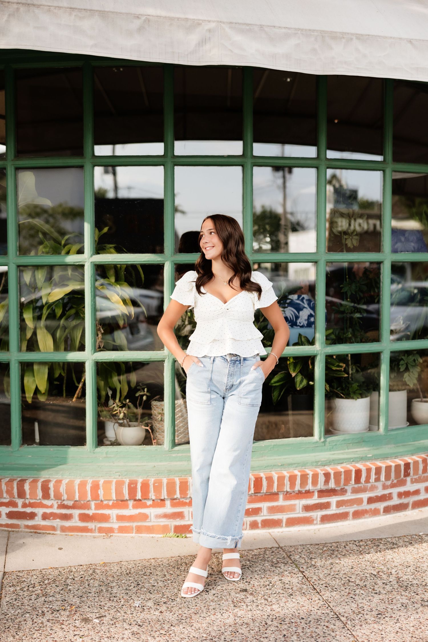 Young woman in white top and light jeans smiling outdoors near a brick storefront with green windows.