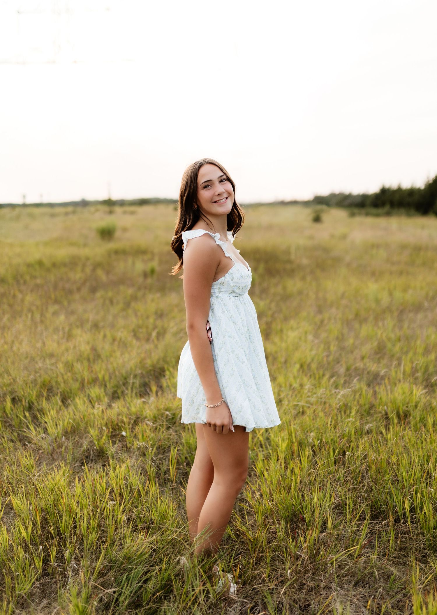 Cheerful young woman in light blue mini dress twirling in open grassy field at sunset.