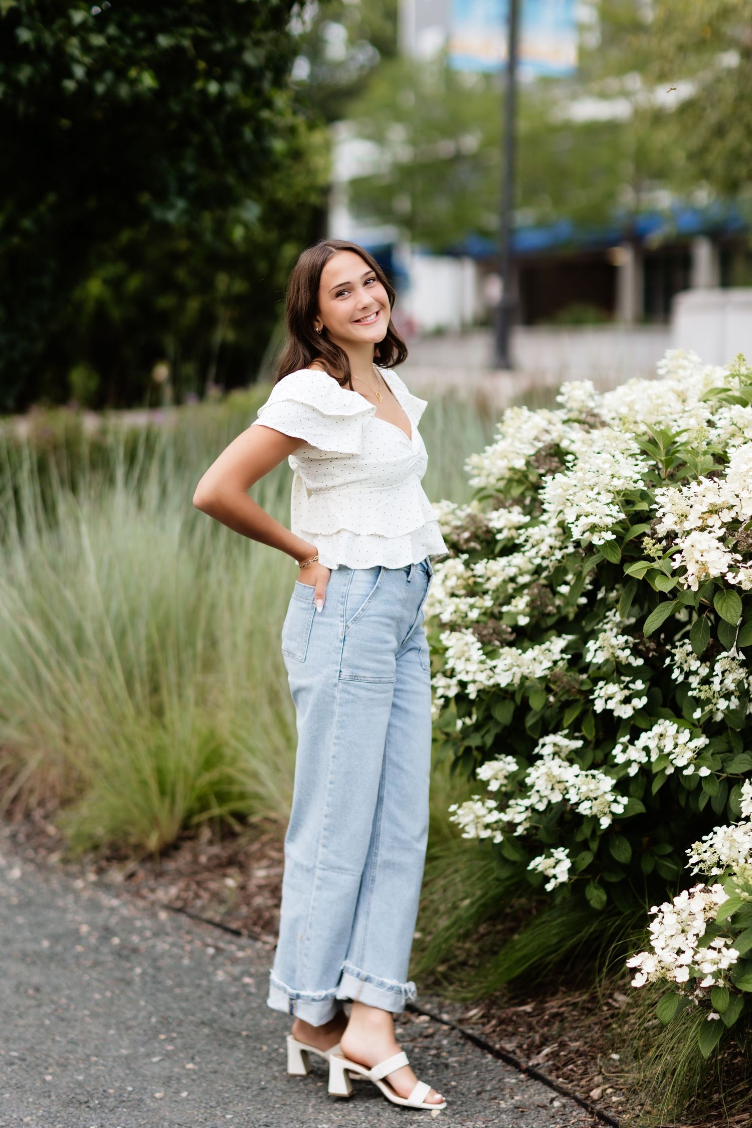 Stylish young woman in white ruffle top and light jeans standing beside lush white flower bushes.