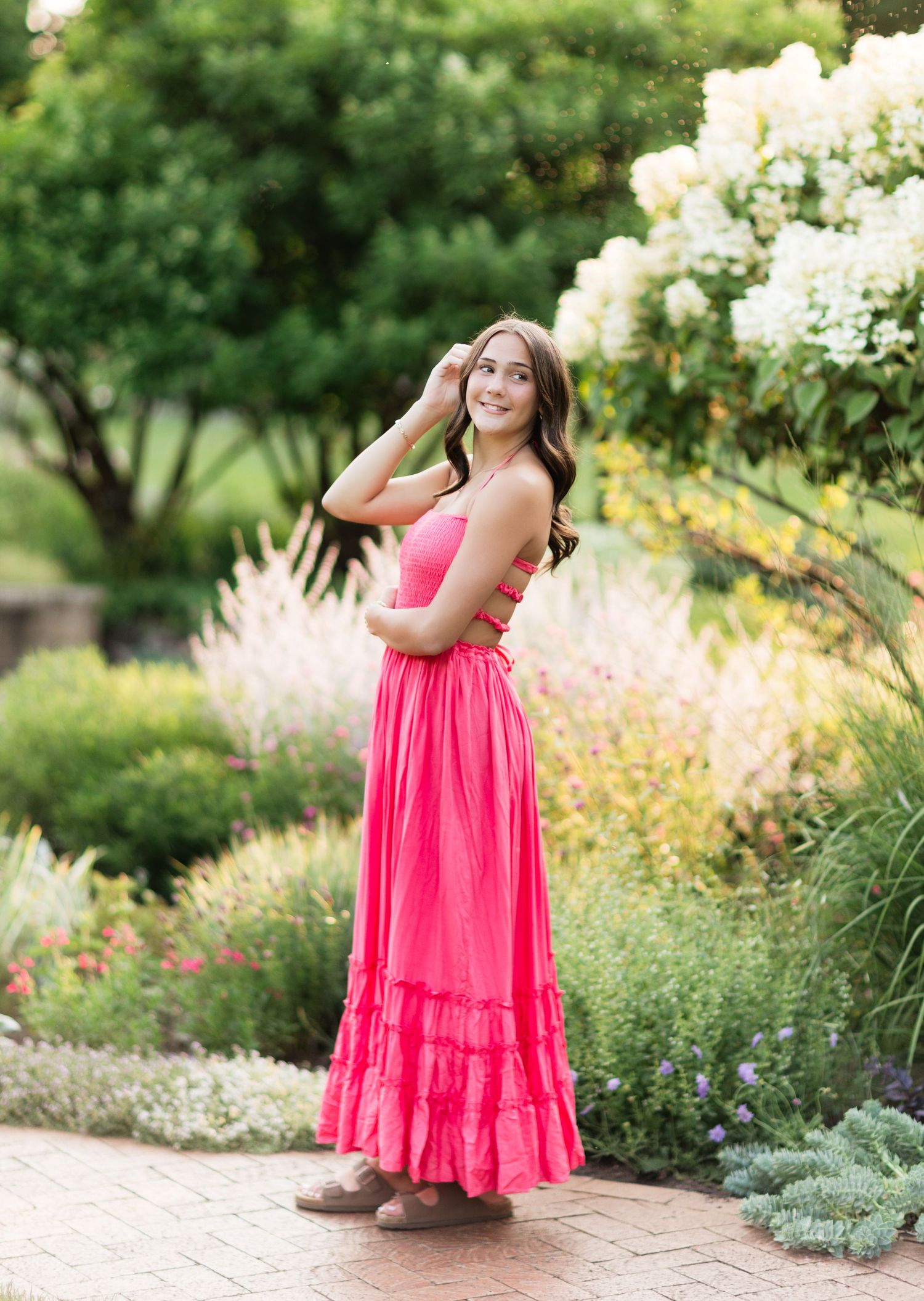Smiling brunette in hot pink maxi dress posing sideways in colorful garden with white flowers.