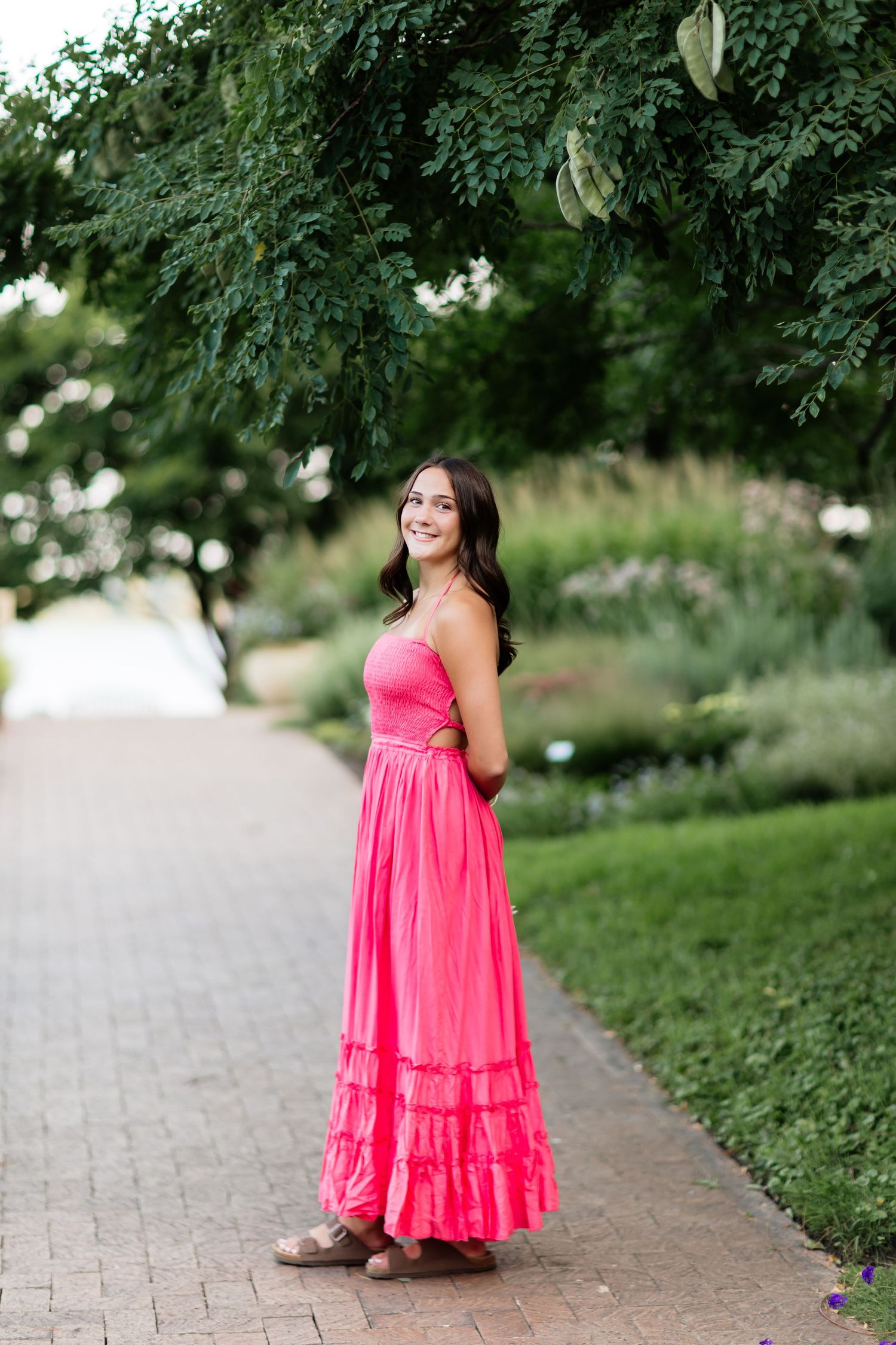 Young woman in a bright pink maxi dress smiling on a brick path surrounded by lush green gardens.