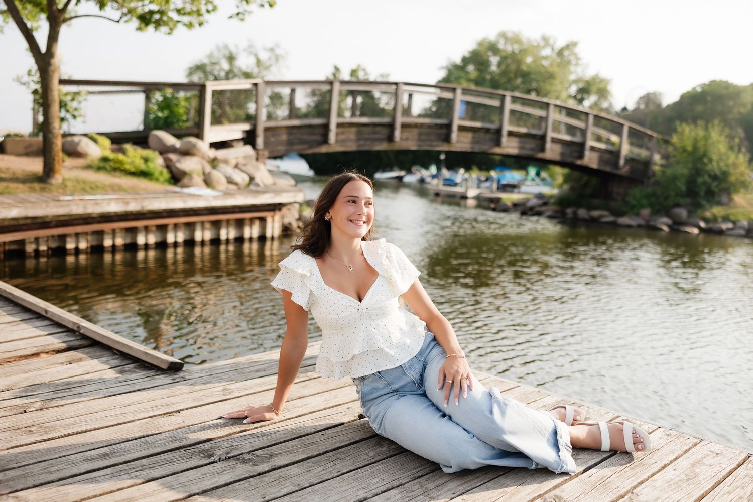 Young woman in white top and jeans sitting on wooden dock by a river bridge, smiling on a sunny day.
