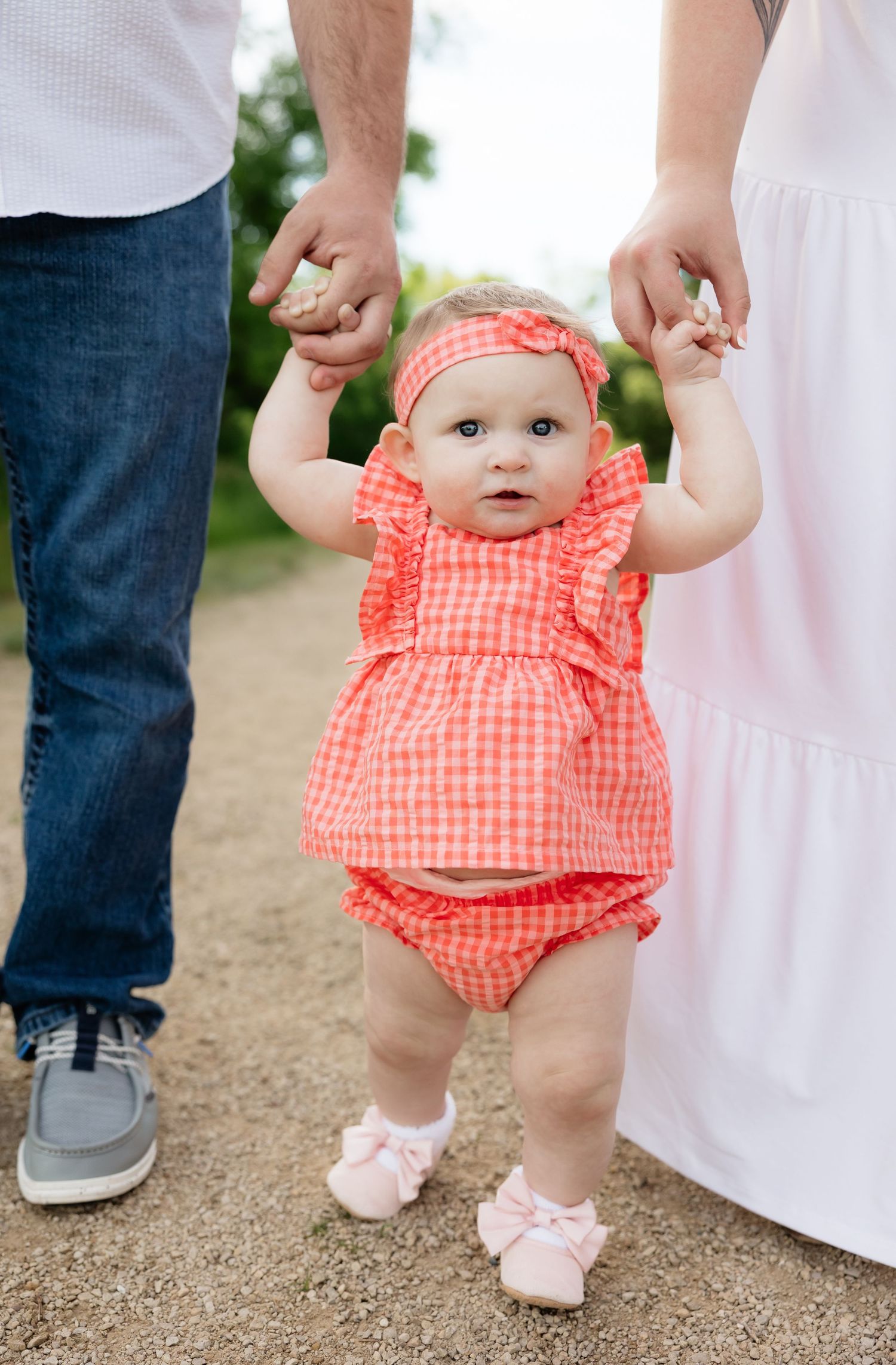 Baby girl in coral gingham outfit held by parents while learning to walk outdoors.