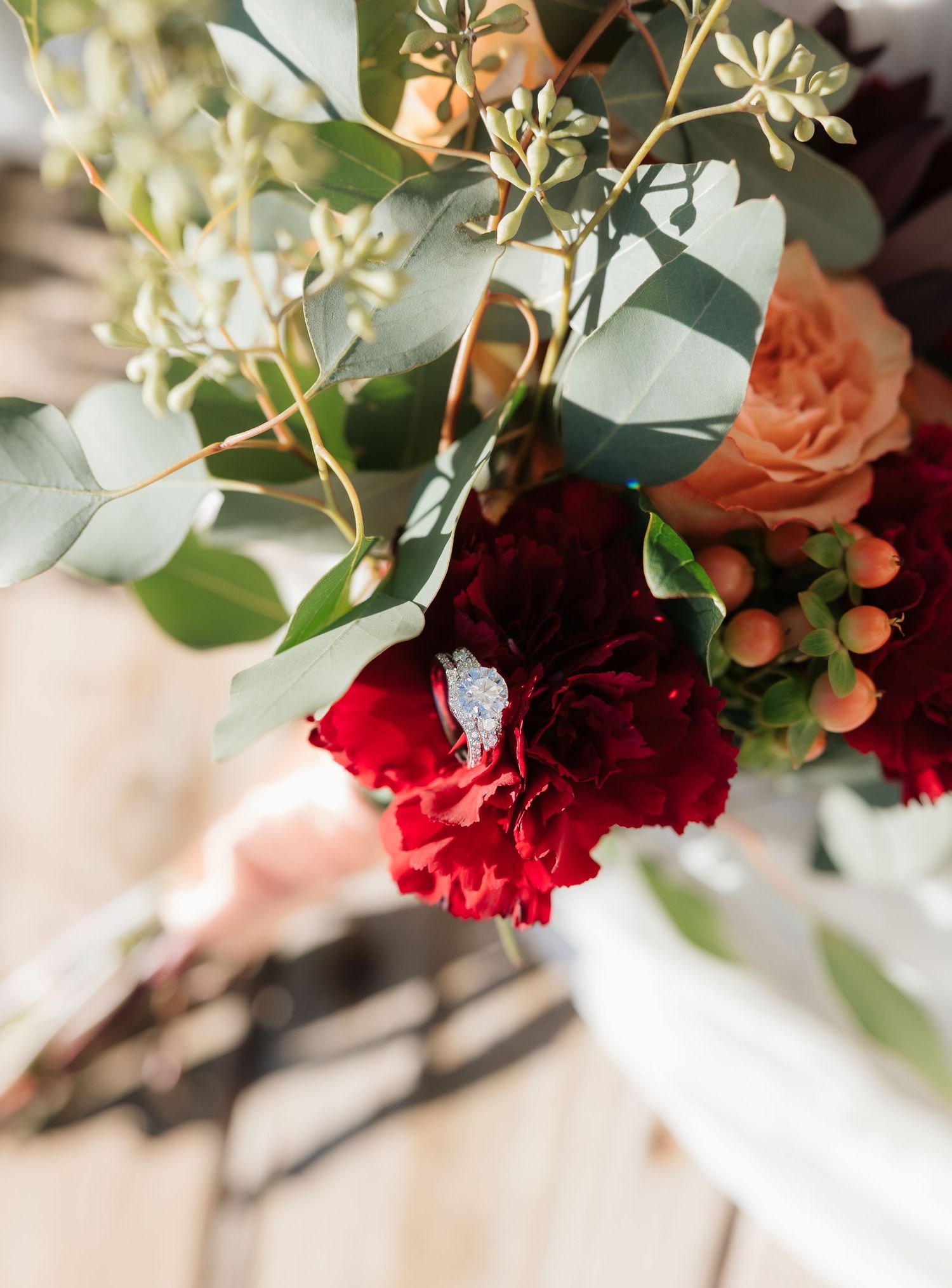 A bridal bouquet featuring deep red carnations, peach roses, eucalyptus, and a diamond engagement ring nestled among the blooms.