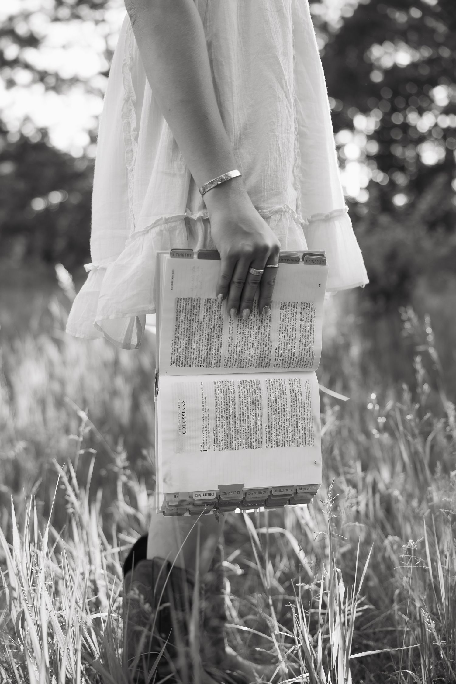 Black and white photo of woman in white dress holding open Bible while standing in tall grass outdoors.