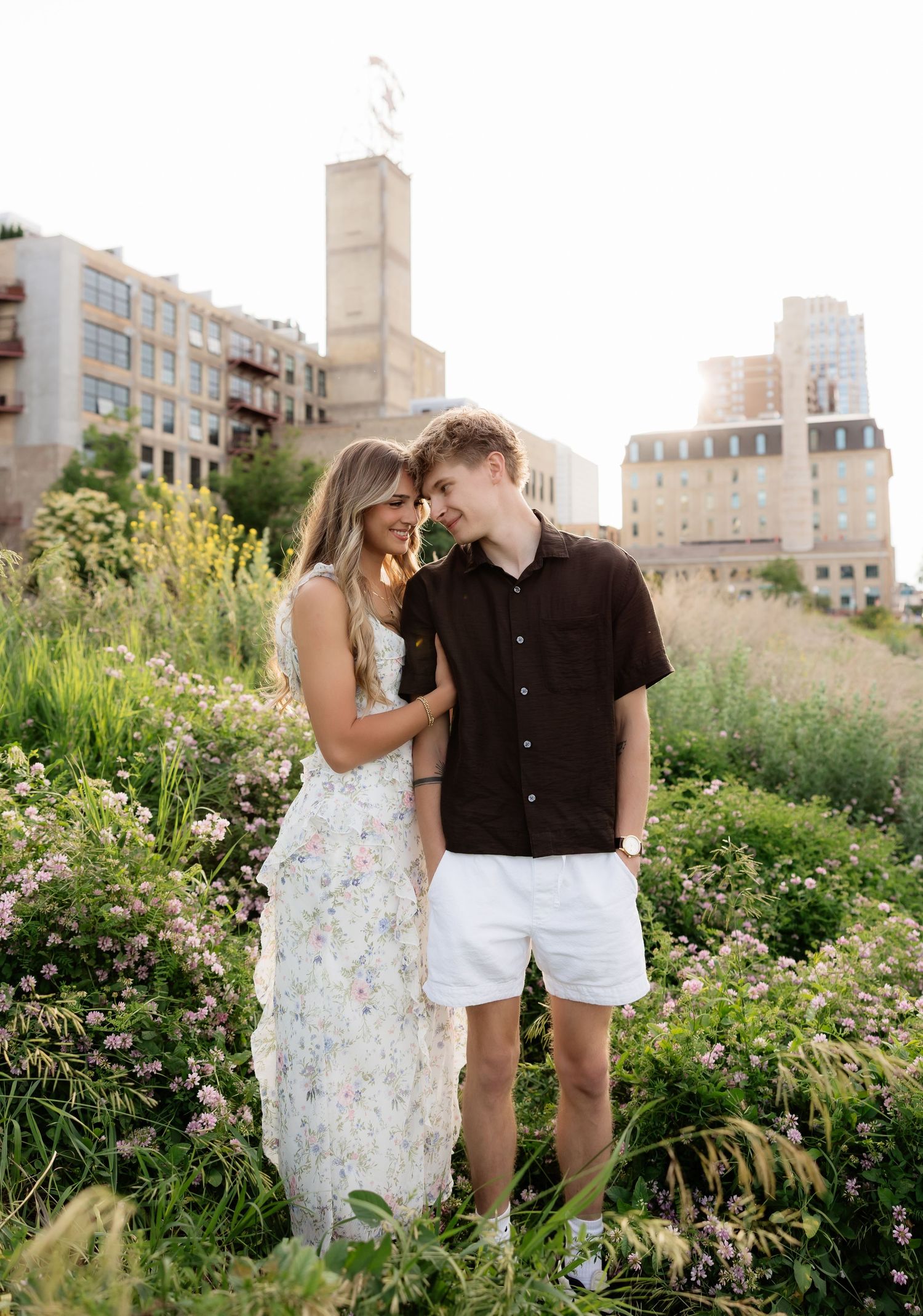 Smiling couple standing close together in wildflower field with urban cityscape glowing behind them.