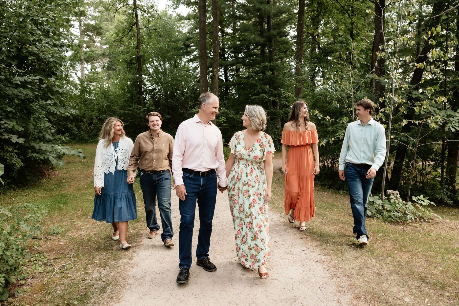 Family of six walks together on a wooded path, smiling and holding hands in coordinated outfits.
