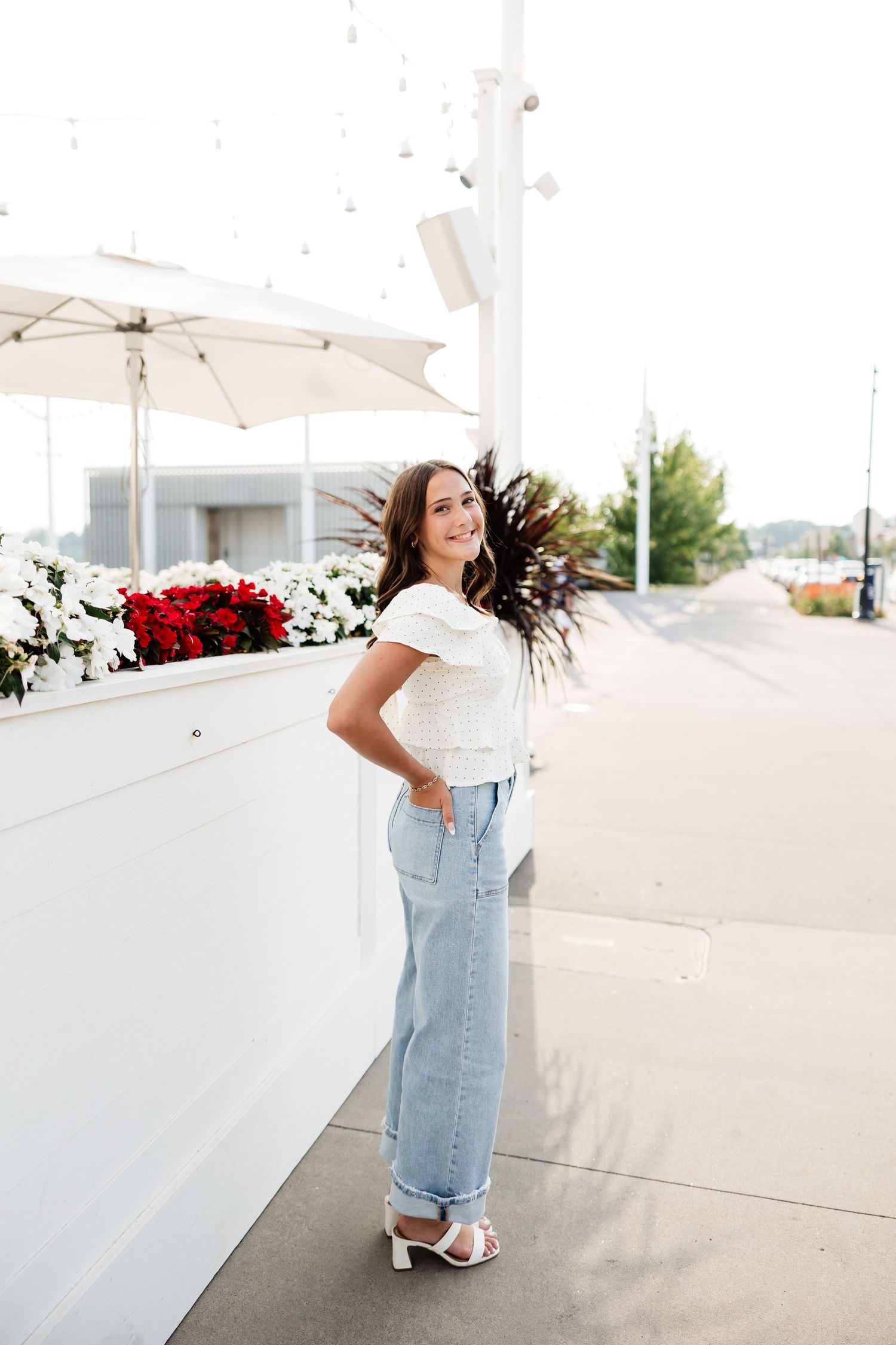 Young woman in white top and wide-leg jeans posing by white wall with red and white flowers.