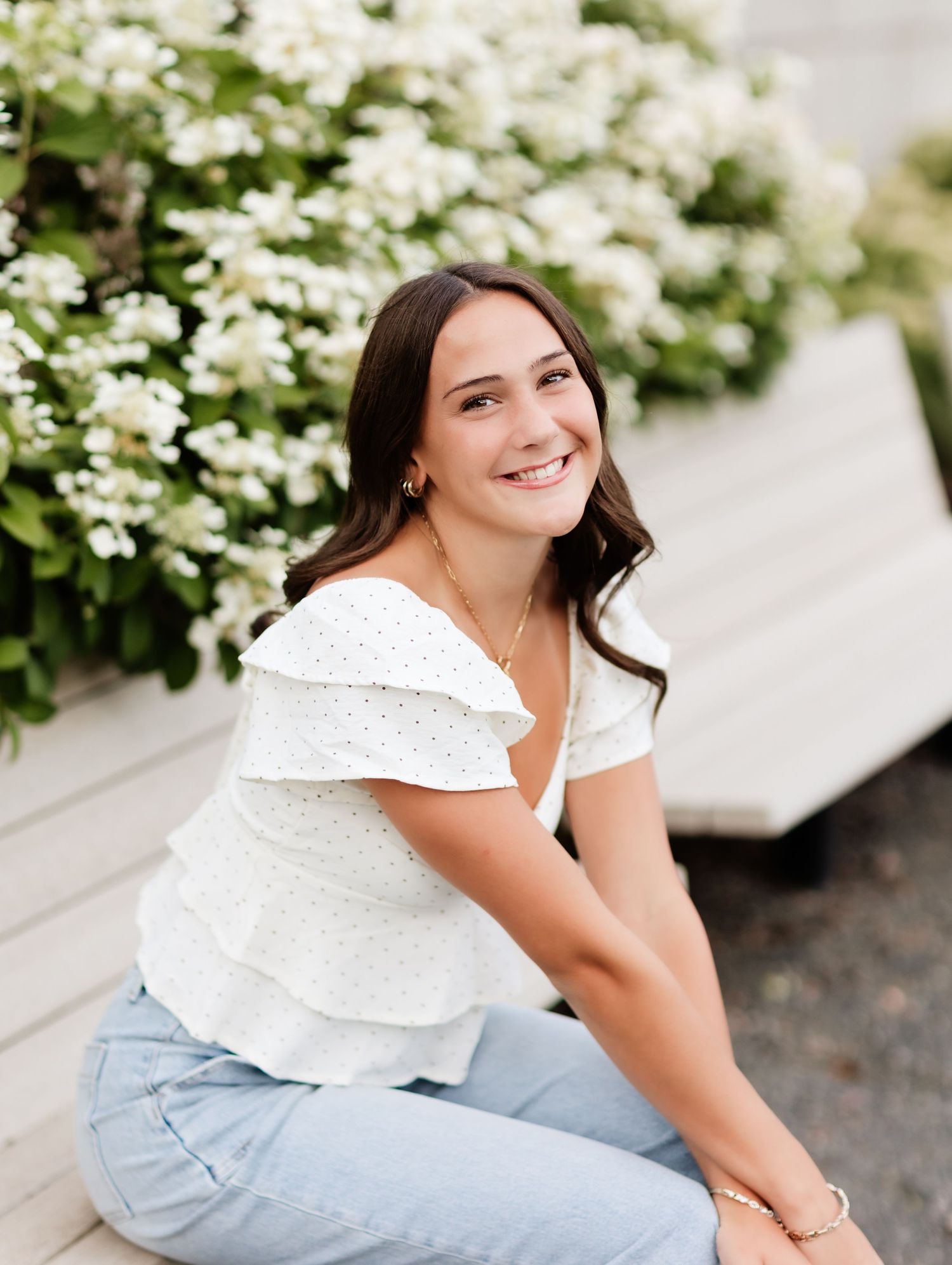 Smiling young woman in white eyelet top and jeans sitting outdoors near white hydrangea flowers.